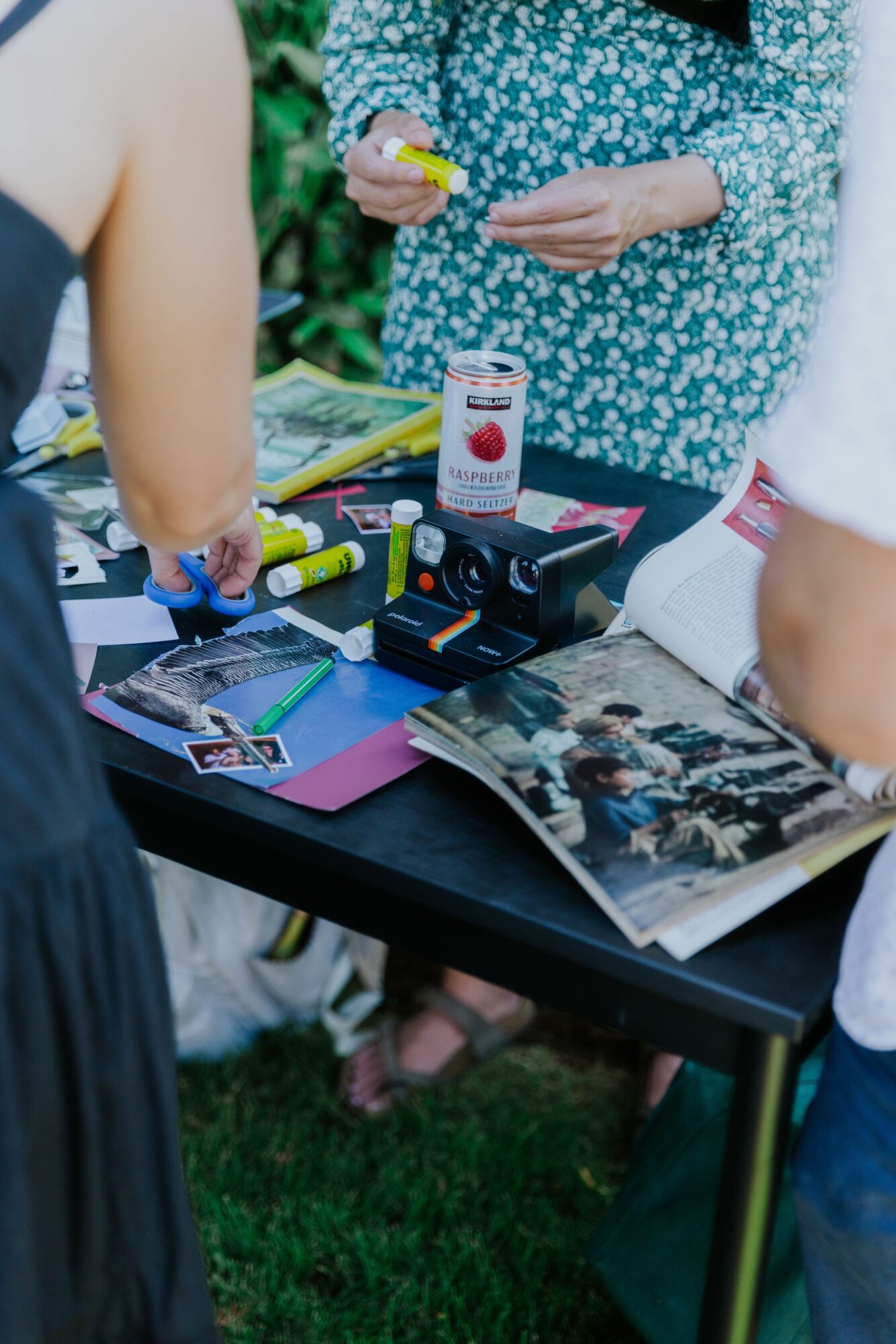 People gathered around a table with cameras, a magazine, and a jar, outdoors with greenery in the background.