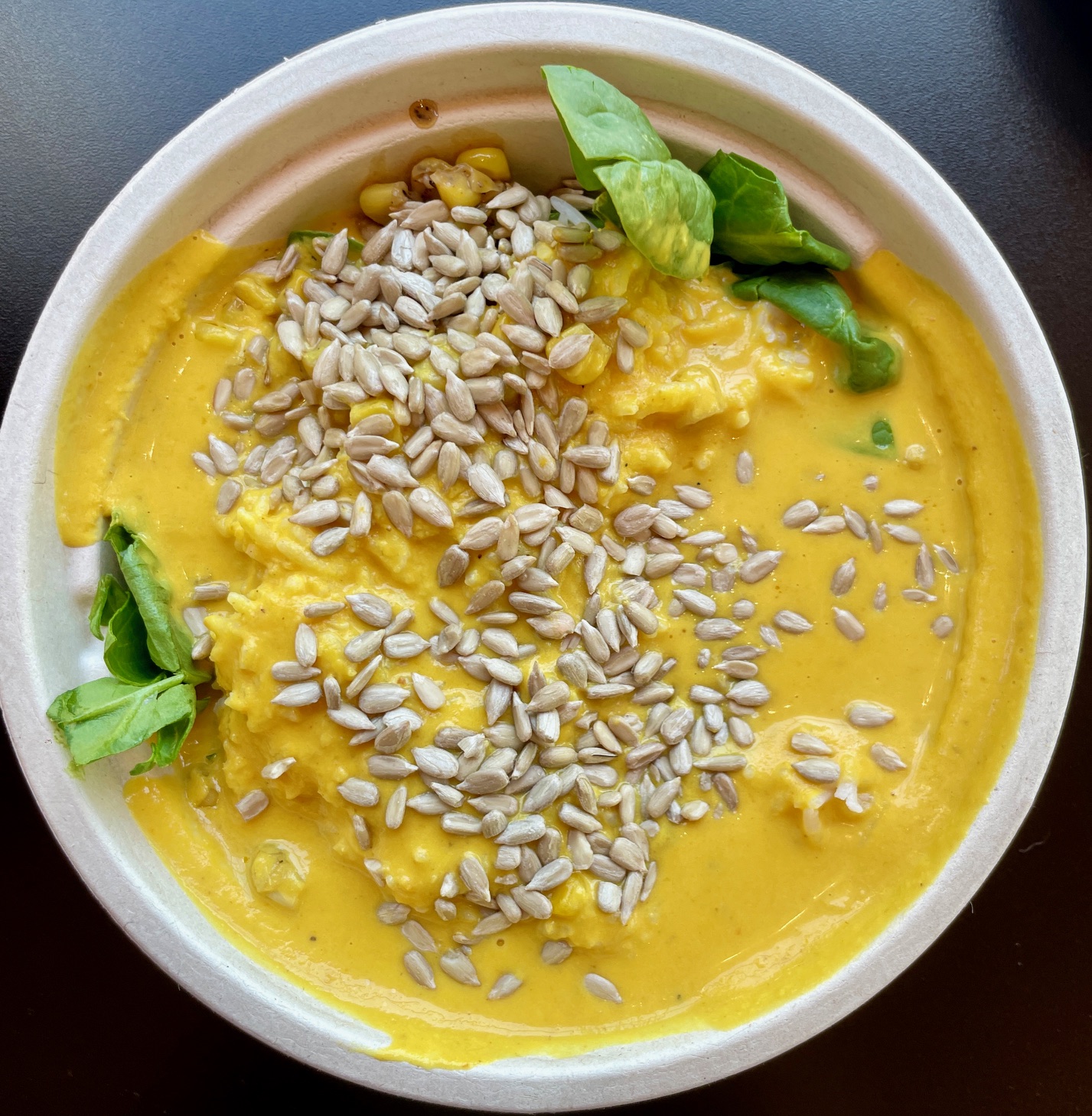 Bowl of yellow soup topped with sunflower seeds and fresh basil leaves, on a dark surface.