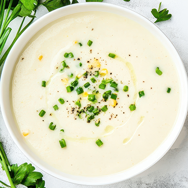 Bowl of creamy soup garnished with chopped green onions and black pepper, with parsley and green herbs around it.