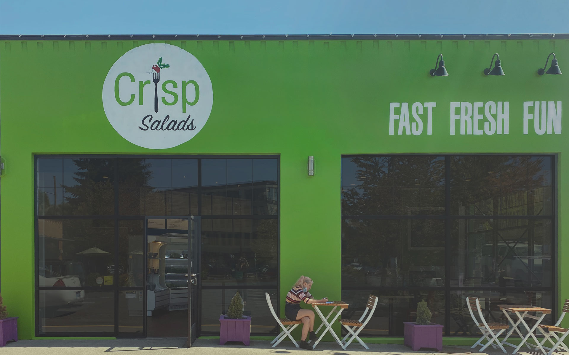 Green storefront with large windows, outdoor seating, and a person sitting at a table, with signage reading 'Crisp Salads' and 'Fast Fresh Fun'.