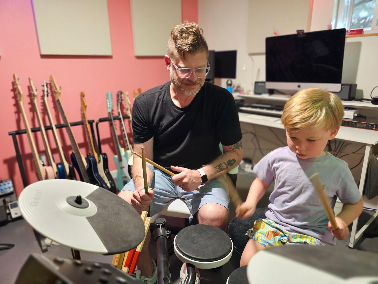 Man and young boy playing drums in a music room with guitars on the wall and computer equipment in the background.