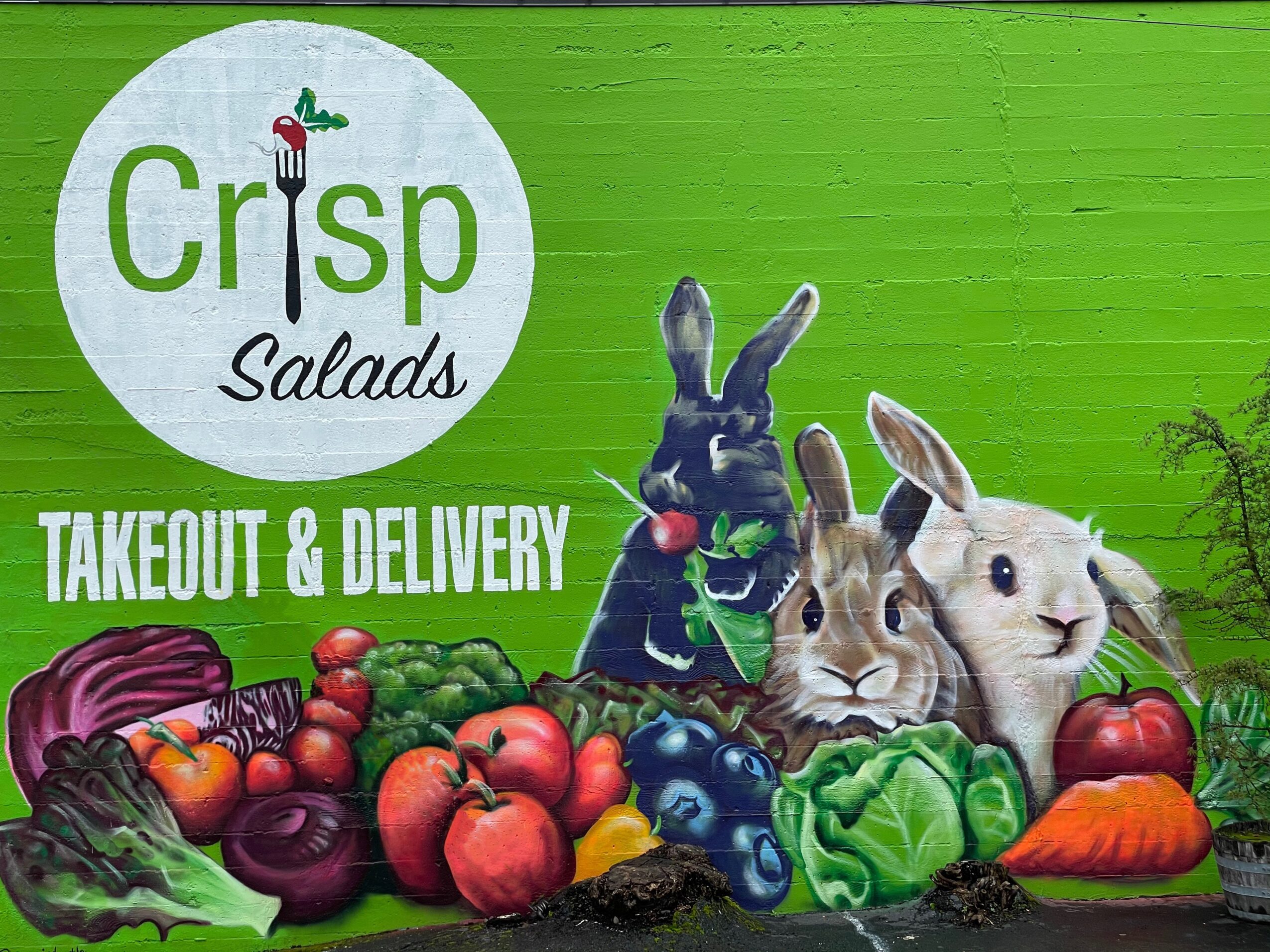 Sign for Crisp Salads with rabbits and vegetables, advertising takeout and delivery, on a green background.