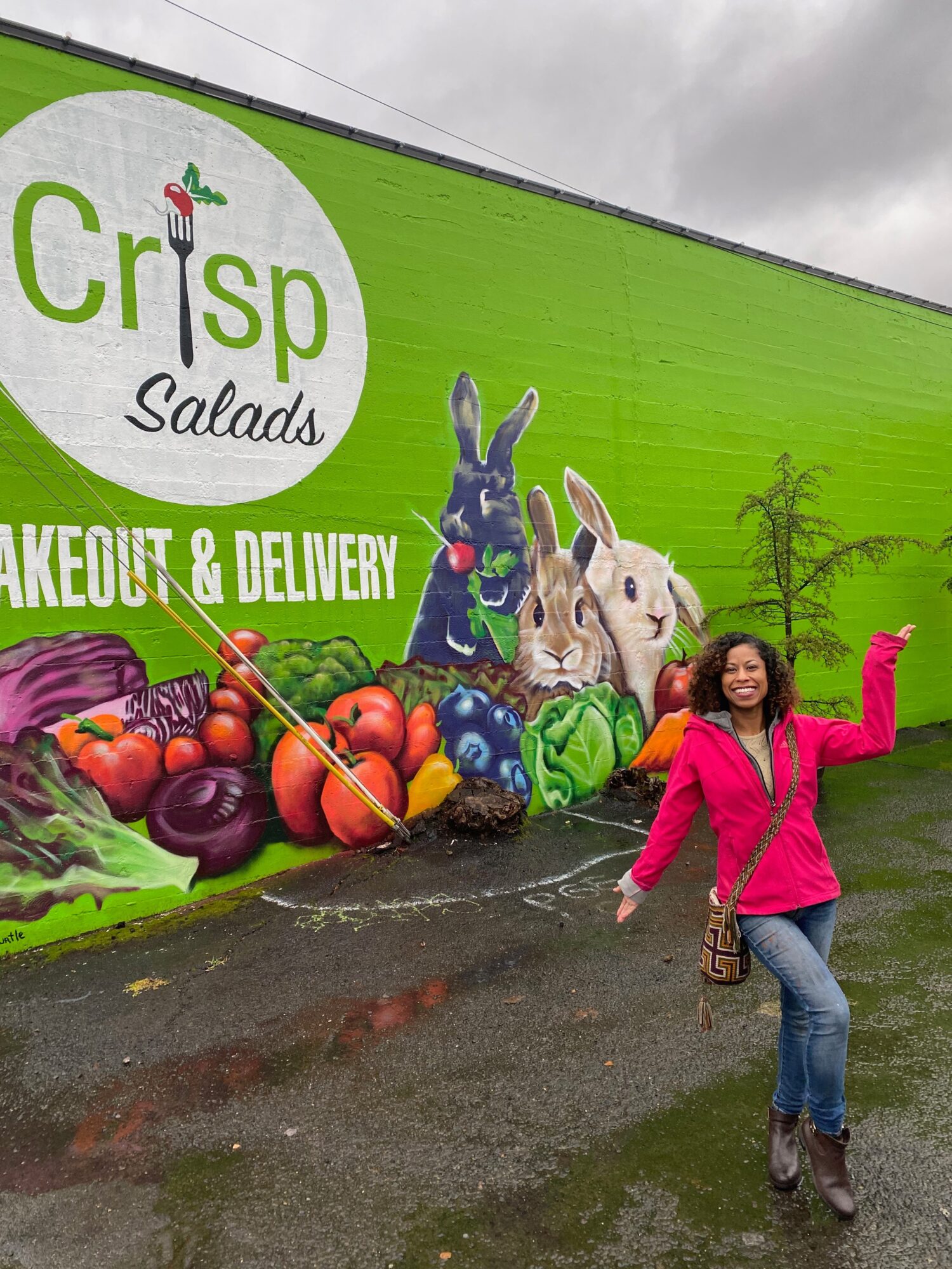 Woman in pink jacket and jeans standing in front of a green mural with rabbits and vegetables, smiling and waving.
