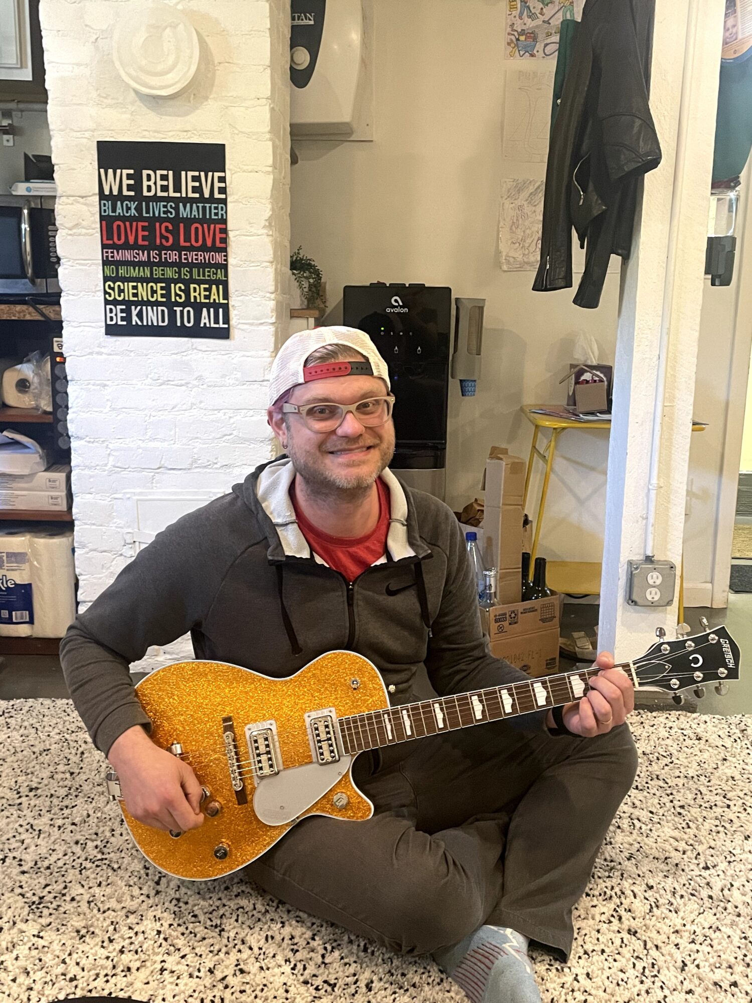 Person sitting on floor holding an orange electric guitar, smiling, in a room with posters and clothing hanging, background clutter.