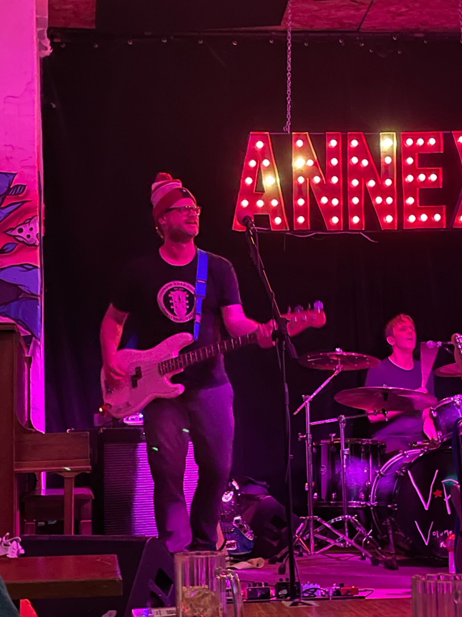 Person on stage holding a guitar, with a band and a lit sign in the background, performing in a dimly lit venue.