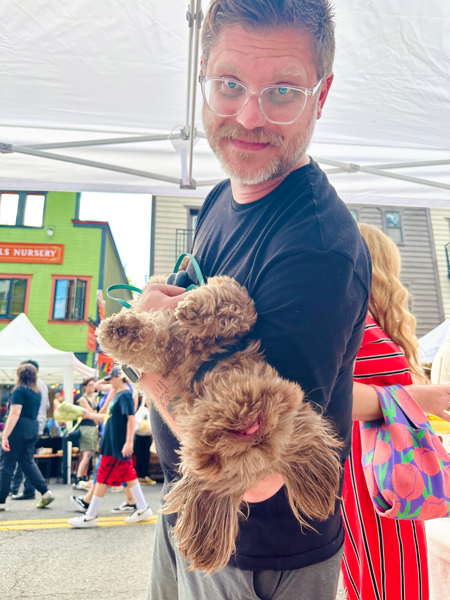 Man holding a fluffy brown dog at an outdoor event with tents and people in the background.