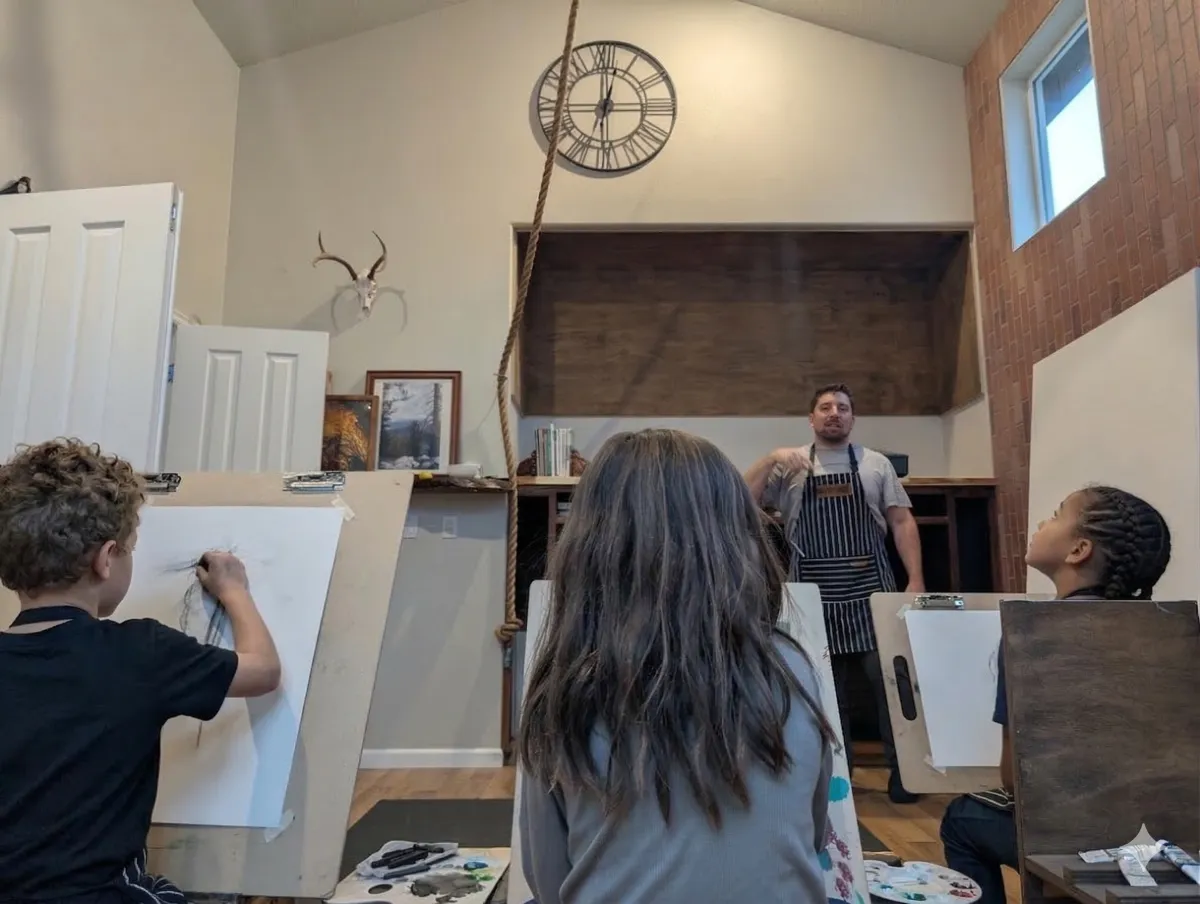 People painting on canvases in an art class, with a teacher at the front, in a room with high ceiling and large window.