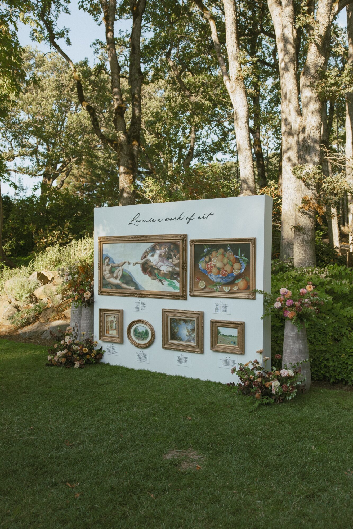Outdoor art display with framed paintings on a white board, surrounded by trees and flowers, sunlight filtering through leaves.