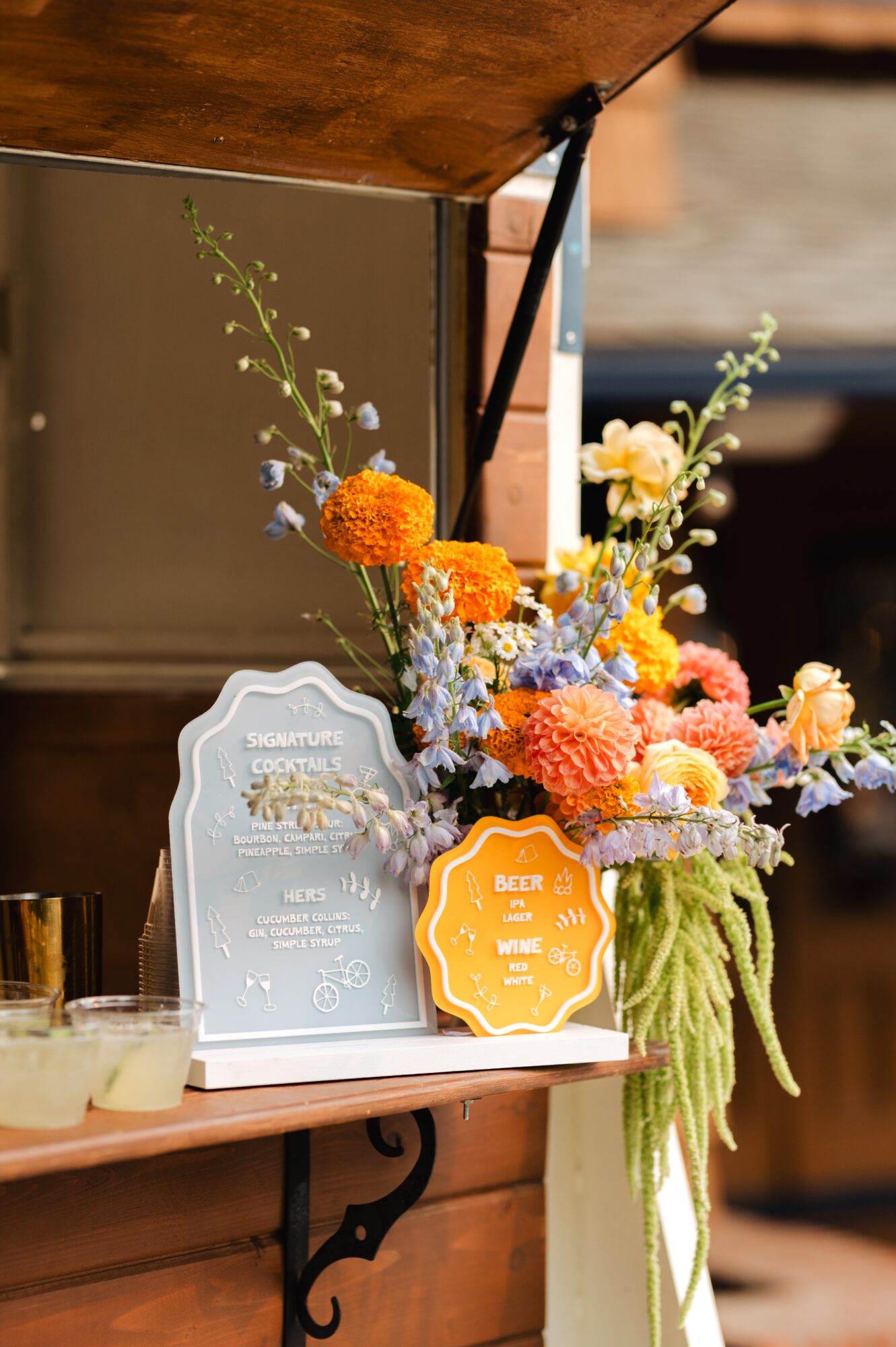 Colorful flower arrangement with orange, pink, and white blooms, and green hanging foliage, on a wooden table with decorative signs.