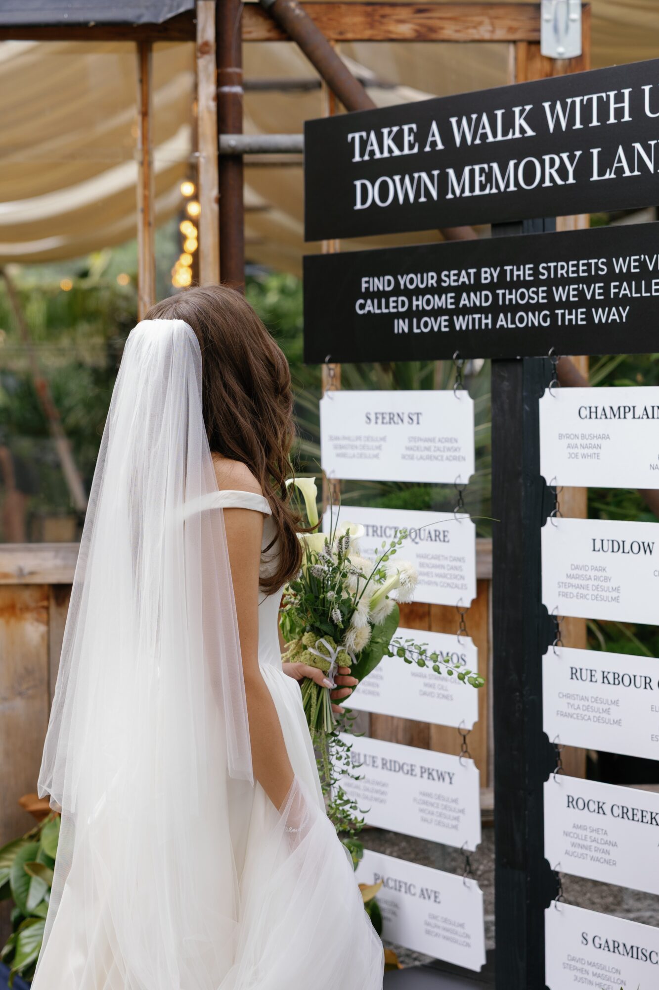 Bride in wedding dress and veil looking at outdoor event signs with flowers and greenery around.