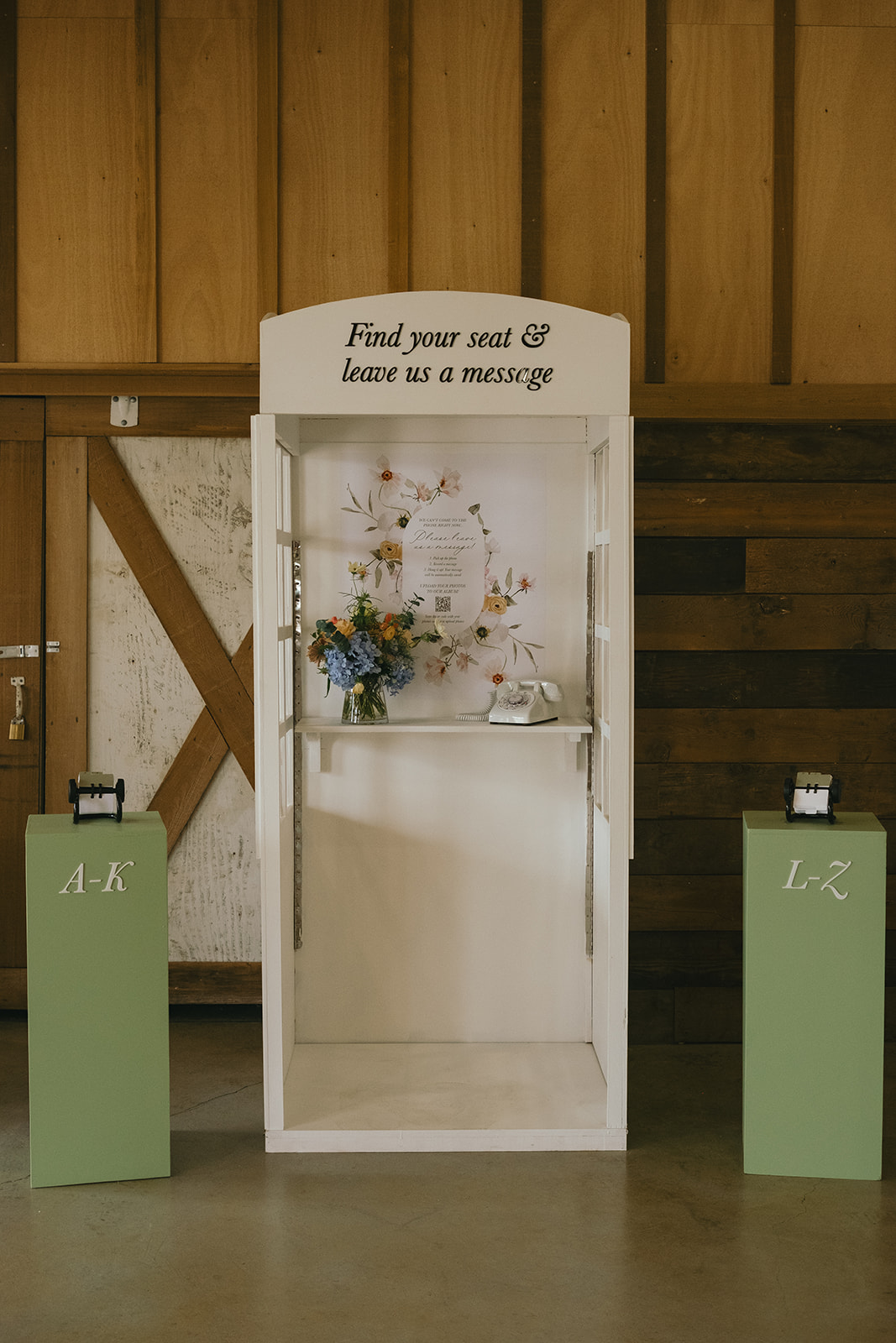 White photo booth with floral decoration, sign on top, and two green and one gray pedestal in front.