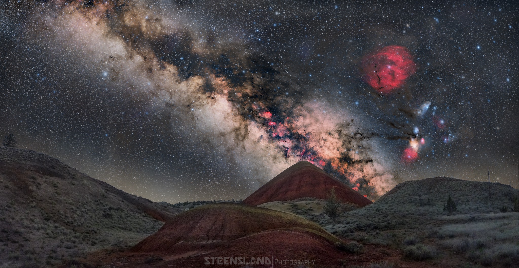 Night sky with the Milky Way galaxy, stars, and colorful nebulae over a landscape with hills.