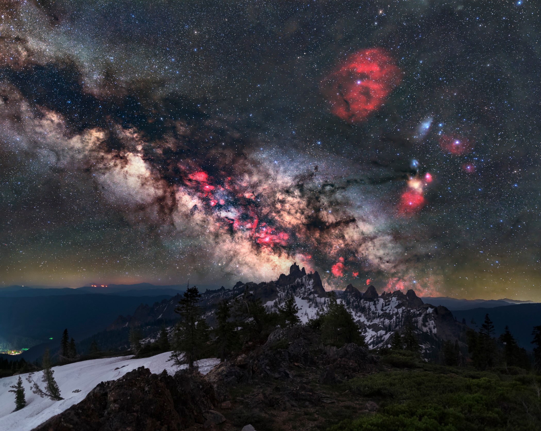 Night sky with stars and colorful nebulae above a landscape with trees and snow, mountain silhouette in the distance.