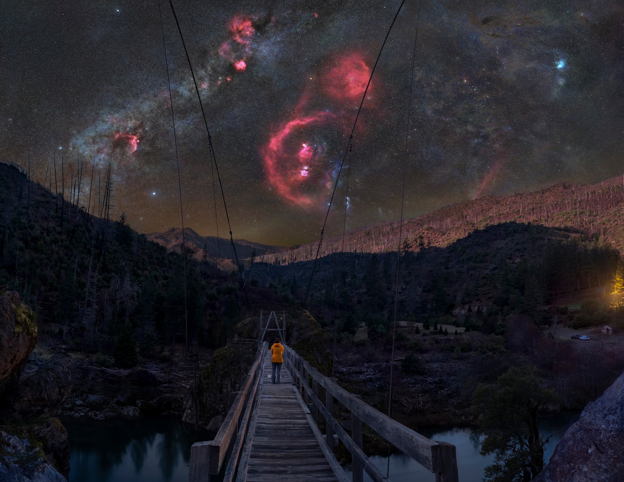 Person standing on a bridge at night with a starry sky and nebulae above, mountains in the background.