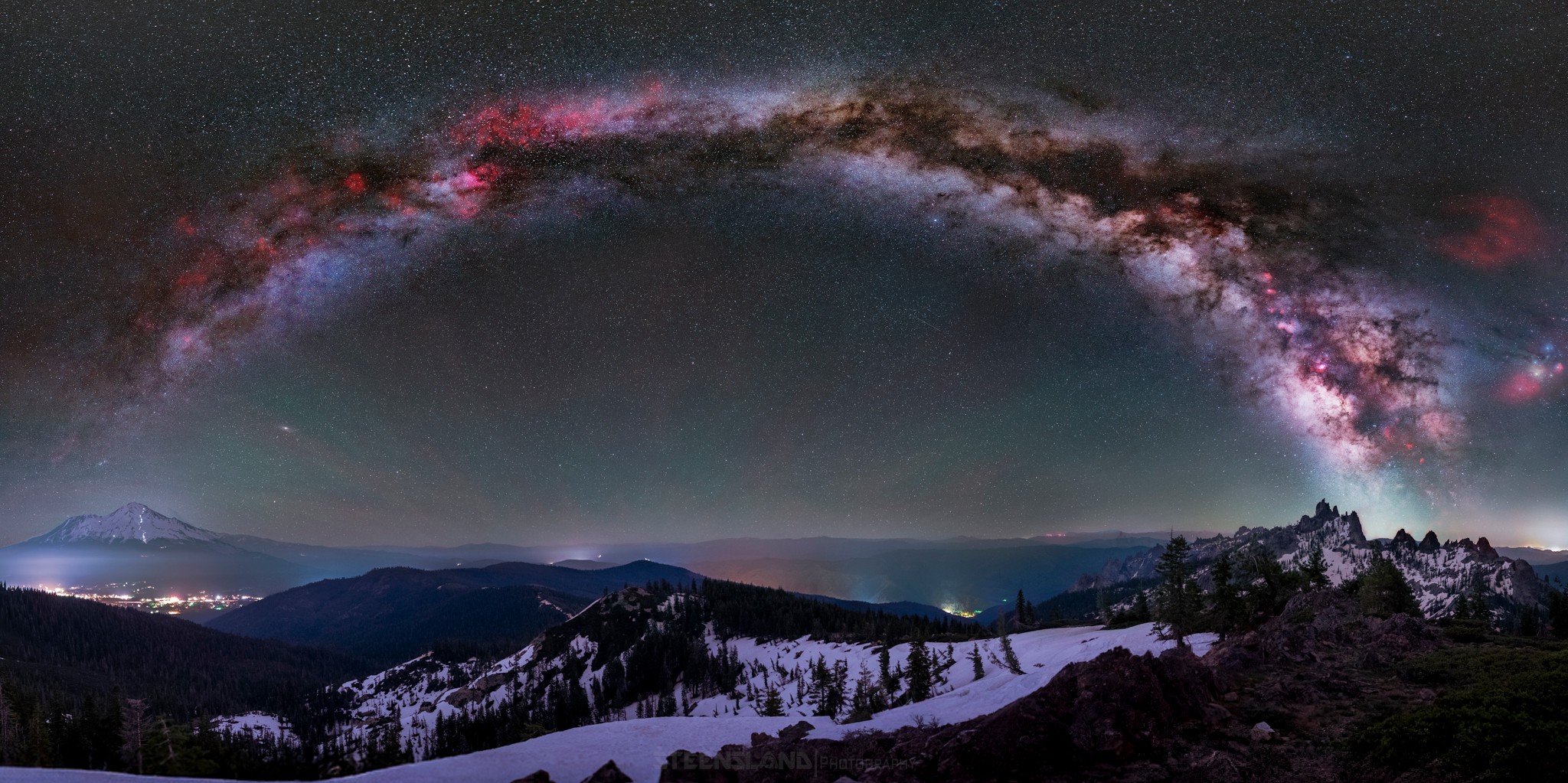 Night sky with a colorful Milky Way arching over snow-covered mountains and trees.