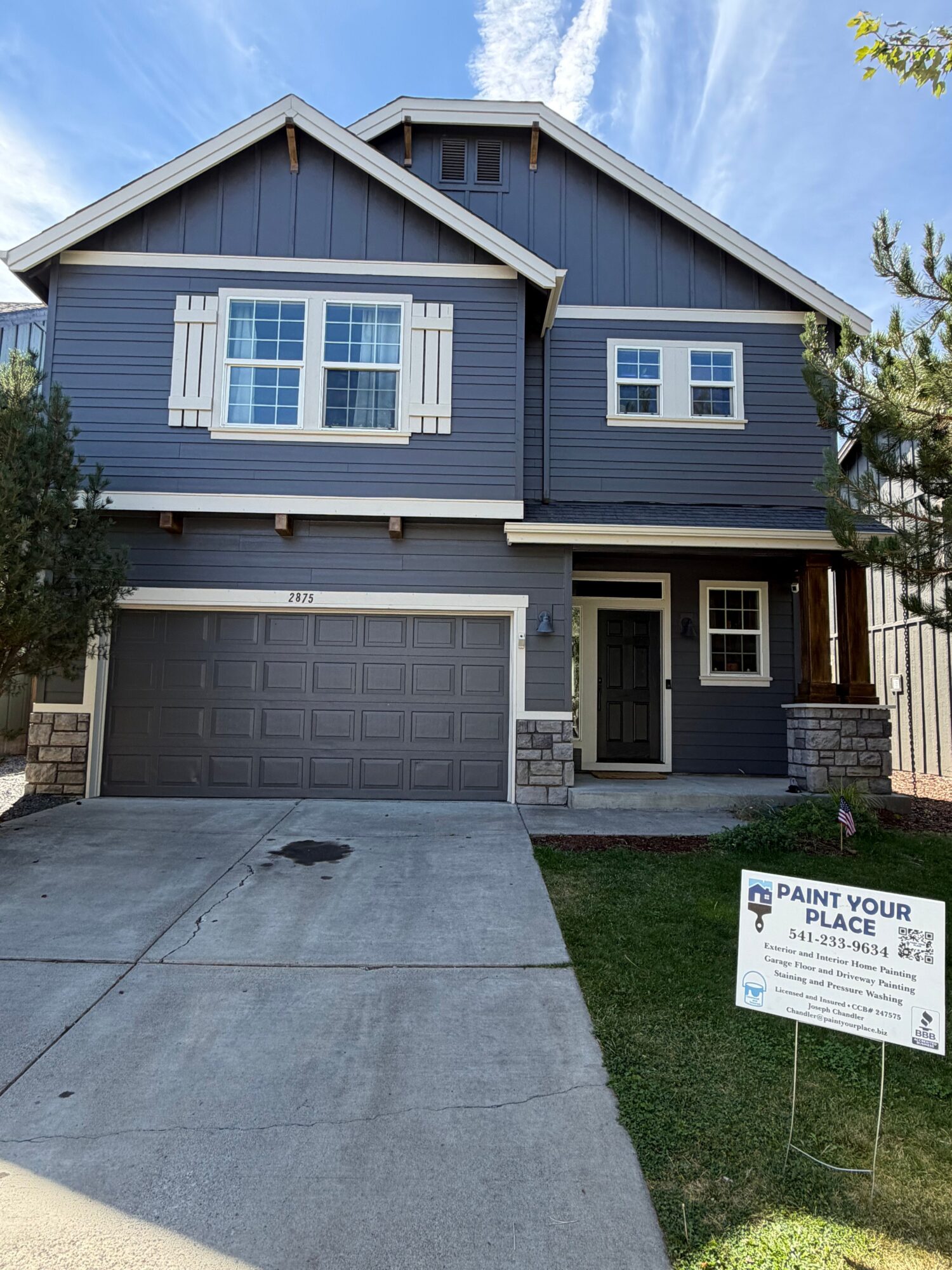 Blue house with white trim, front door, garage, concrete driveway, and a sign in the yard, under a blue sky.