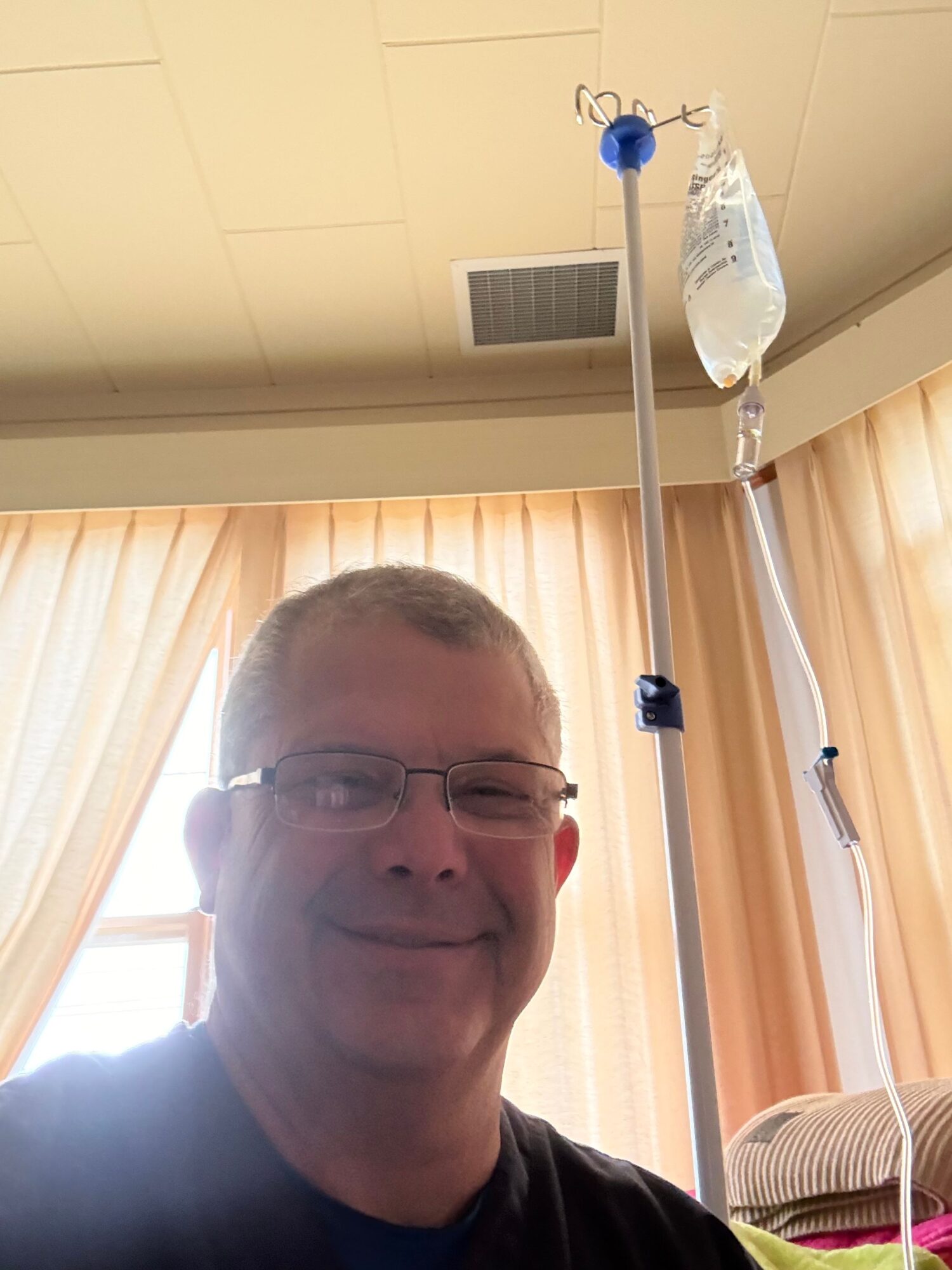 Person with glasses and short hair in a room with beige curtains and medical equipment.