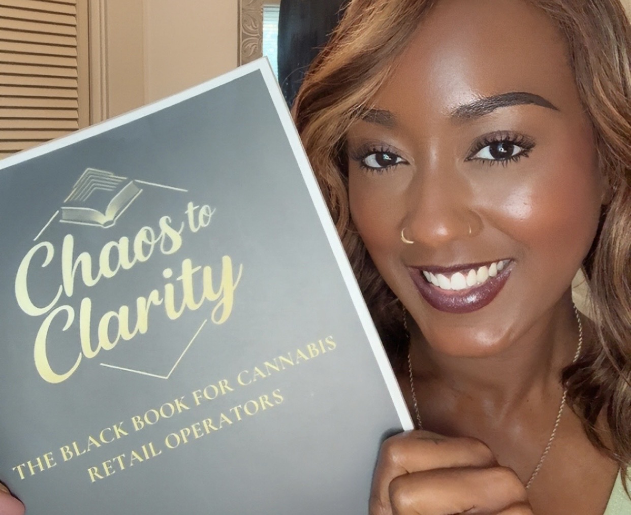 Woman with curly hair smiling holding a book titled 'Chaos to Clarity' in a room.