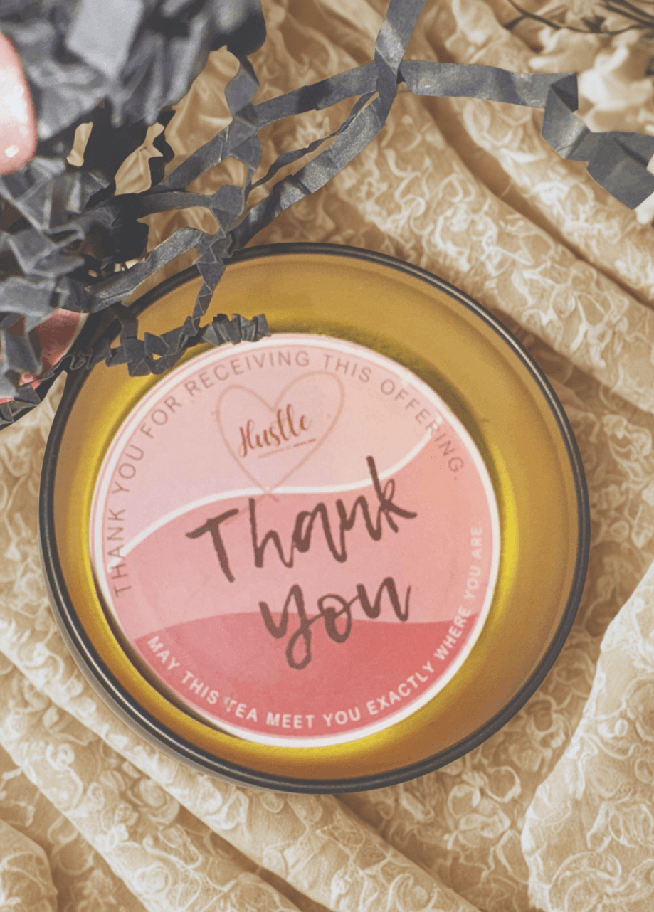 Round yellow container with a pink and white label reading 'Thank you' and 'Hustle', placed on crumpled beige fabric.