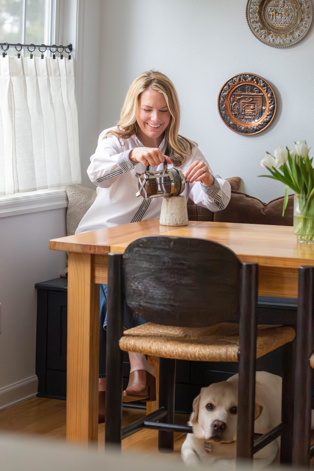Woman smiling at table, dog lying under chair, flowers on table, decorative plates on wall, sunlight through window.