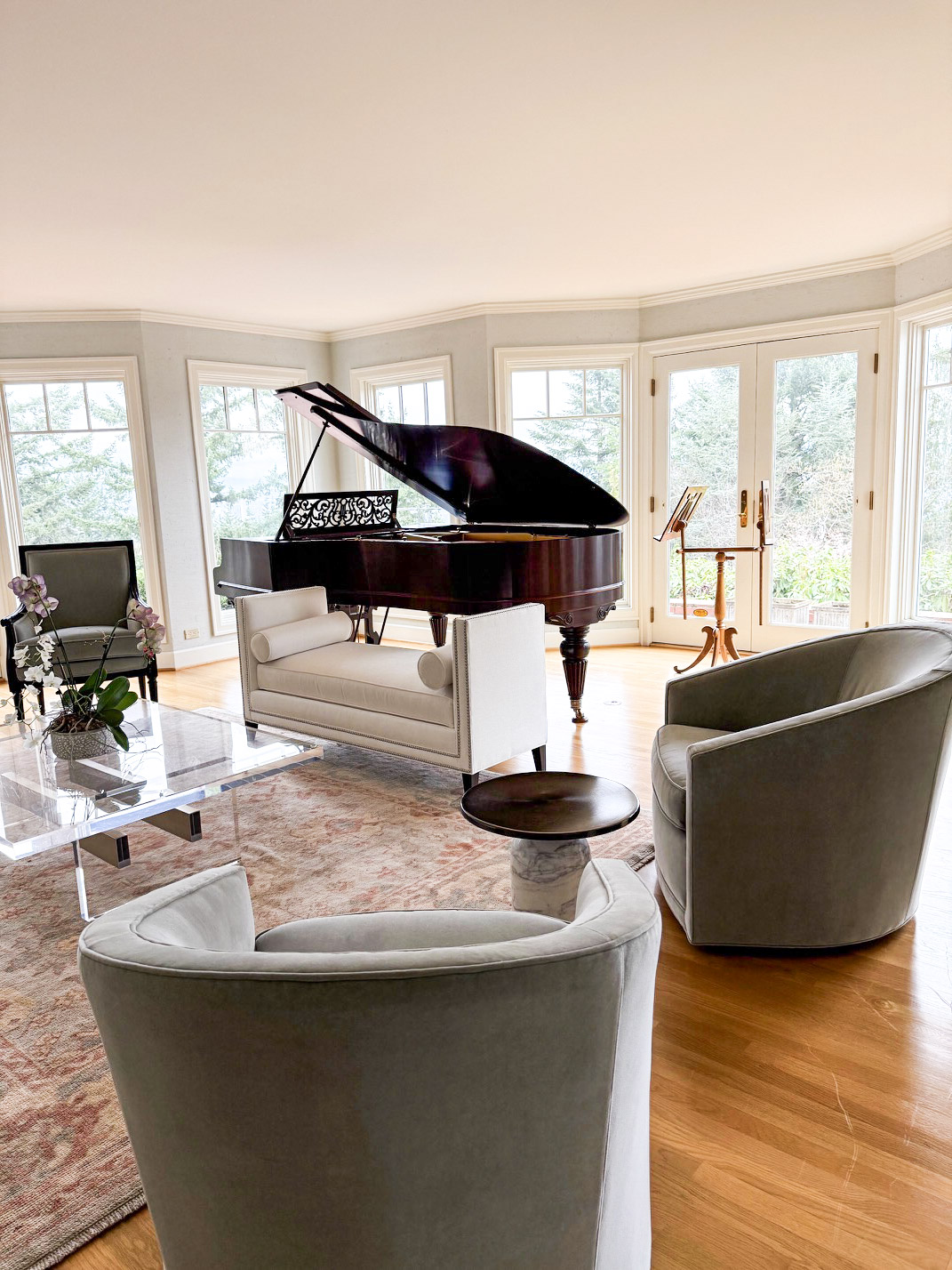 Living room with grand piano, two armchairs, a glass table, and large windows with a view outside.