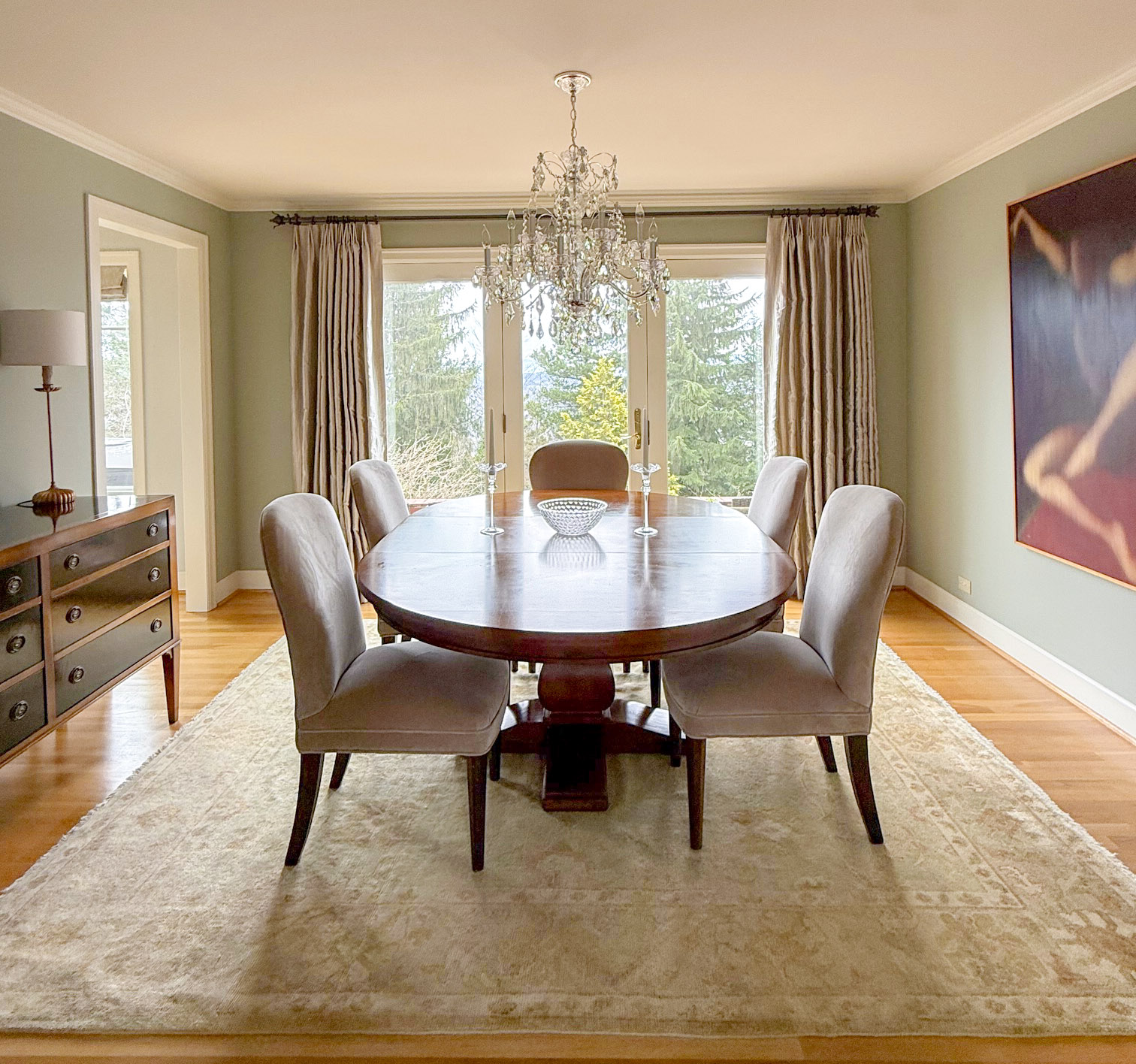 Dining room with round table, six chairs, chandelier, large window with curtains, and artwork on wall.