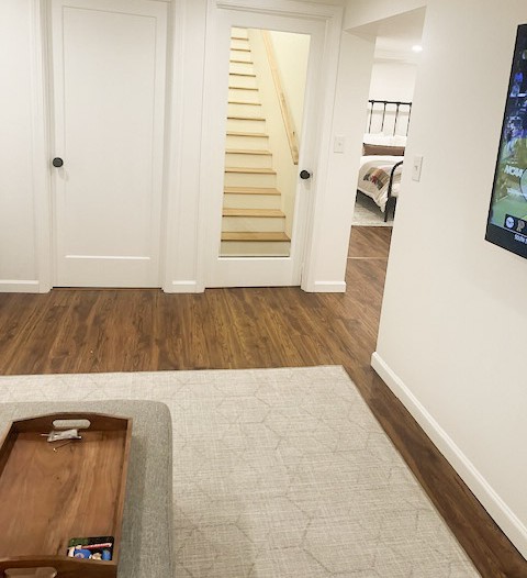 Living room with hardwood floor, white walls, and a staircase visible through an open door, with part of a bed in a bedroom.