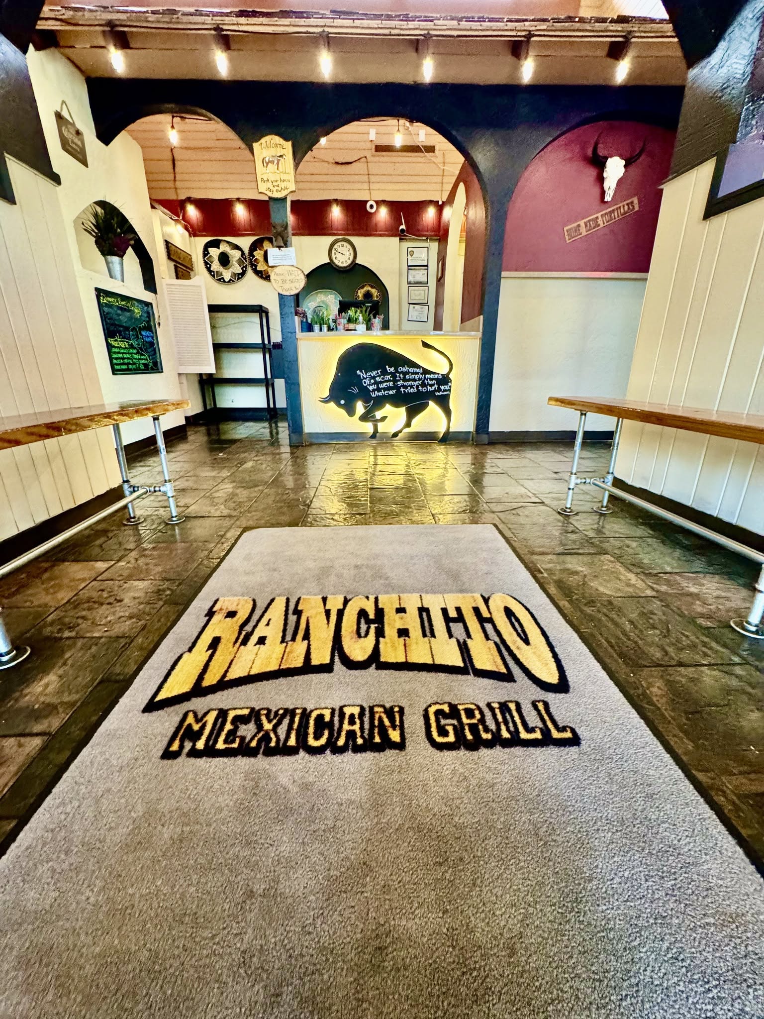 Entrance area of a Mexican grill restaurant with a branded mat, wooden floors, and colorful wall decorations.