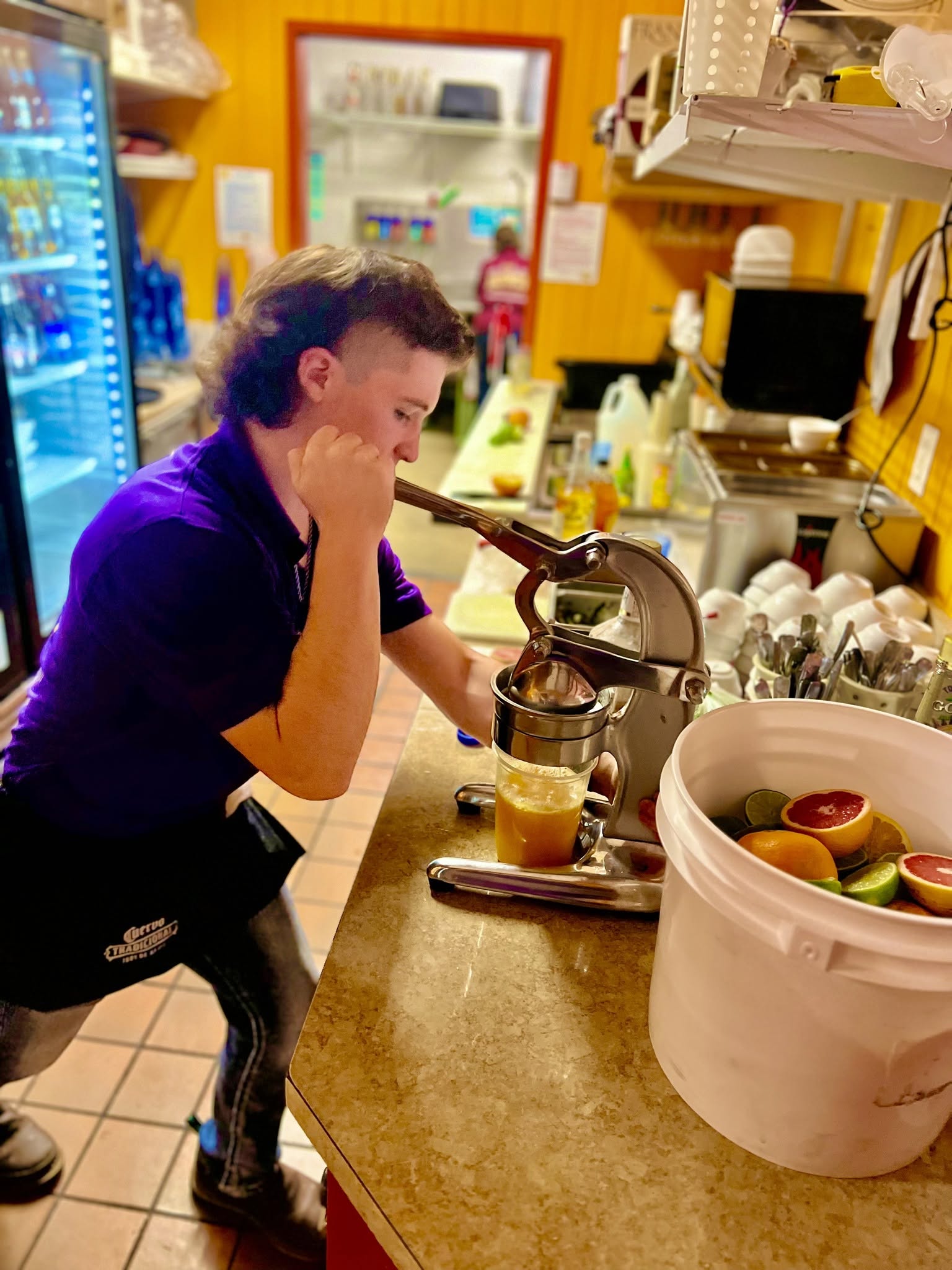 Young person with curly hair in a purple shirt stands at a coffee machine, preparing a drink in a colorful kitchen.