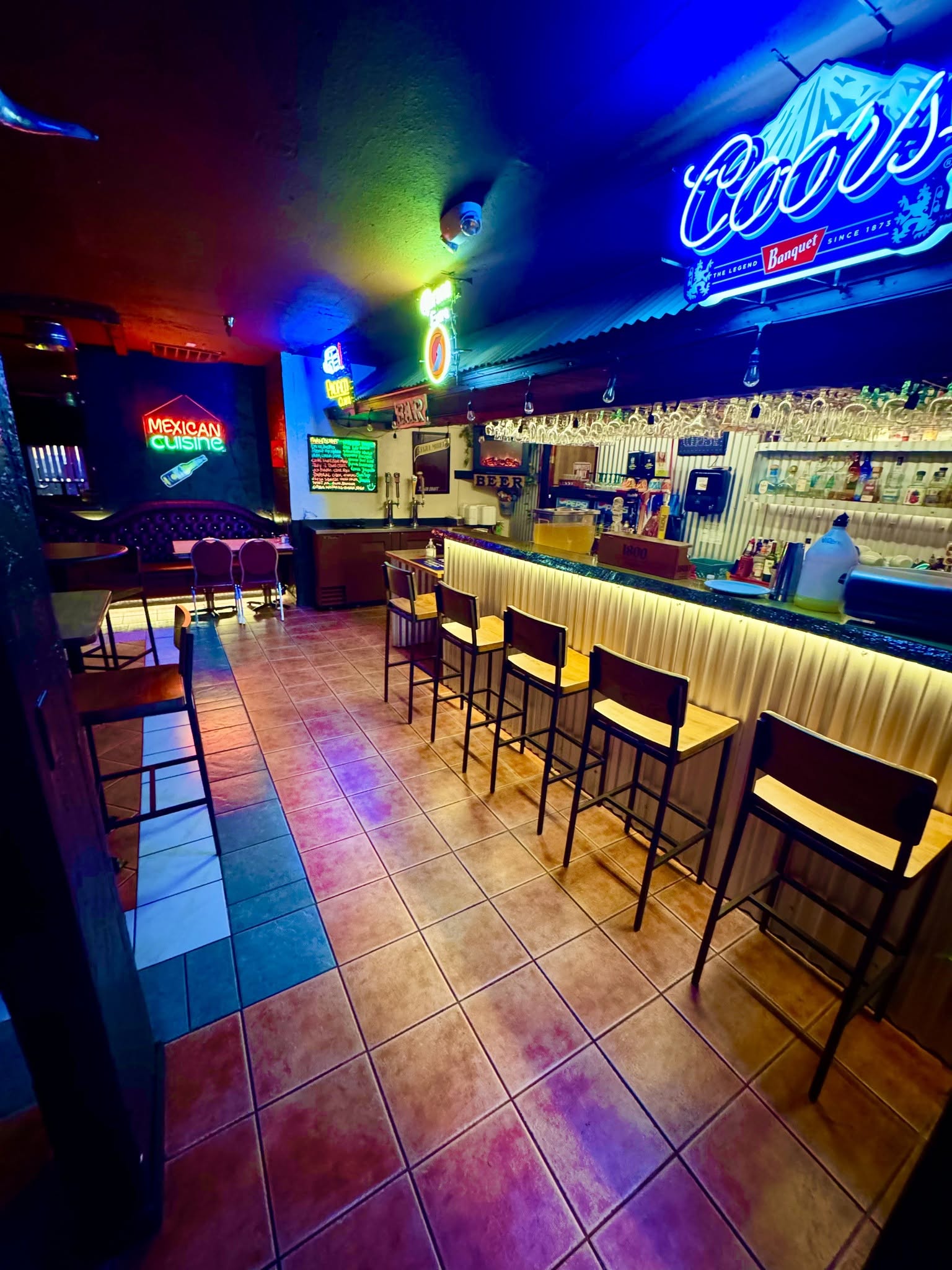 Bar with illuminated neon signs, colorful lighting, and a row of chairs along the counter, with shelves of bottles behind the bar.