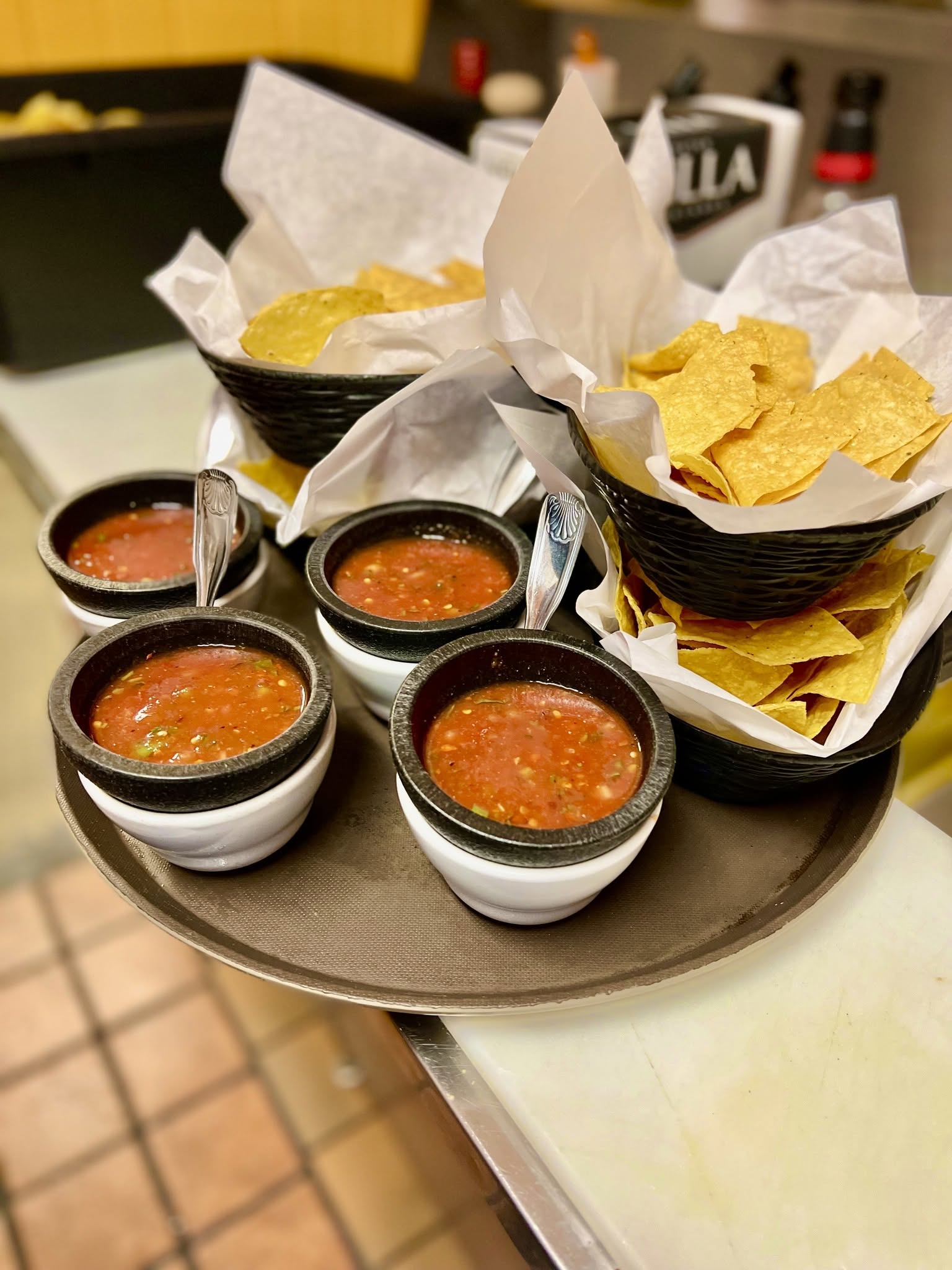 Baskets of tortilla chips with bowls of salsa on a black tray, with a napkin-wrapped item in the background.