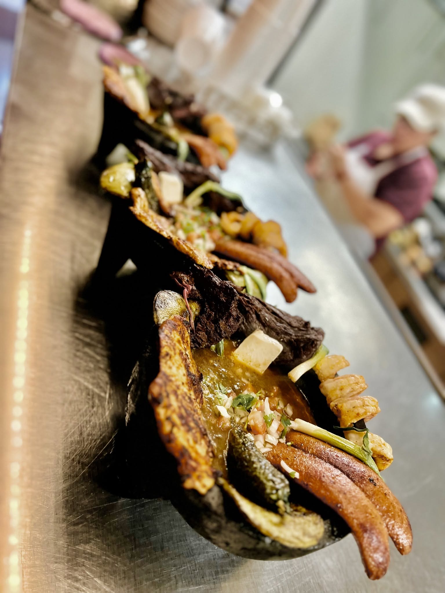 Close-up of a row of stuffed eggplants on a metal surface, with a person in the background holding food, blurred.