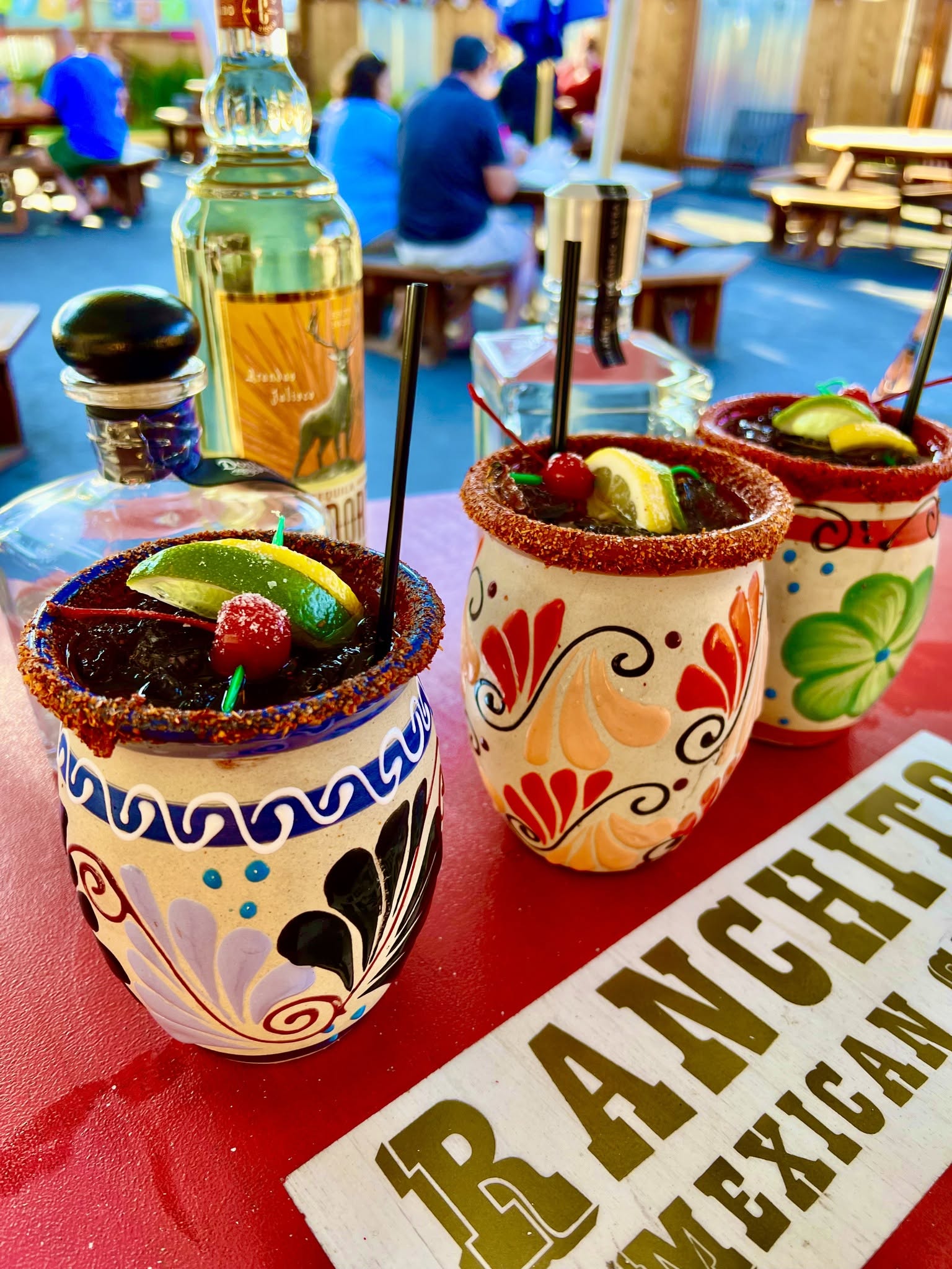 Three colorful margarita glasses with fruit garnishes on a table, with a lively restaurant scene in the background.