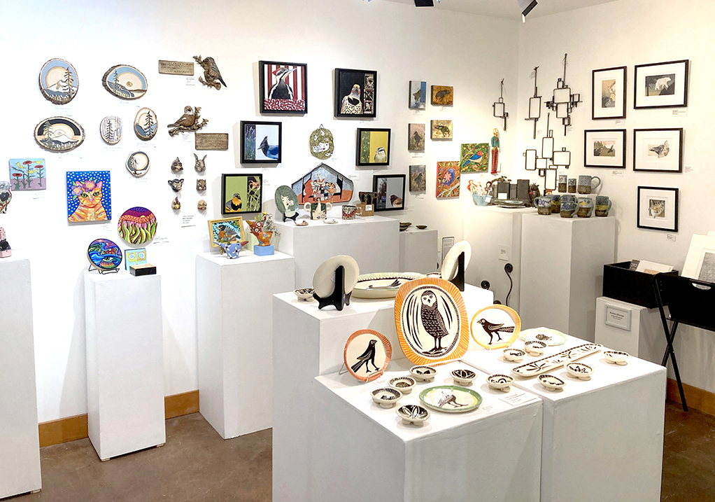 Art gallery with paintings, ceramics, and decorative objects displayed on white pedestals and walls.