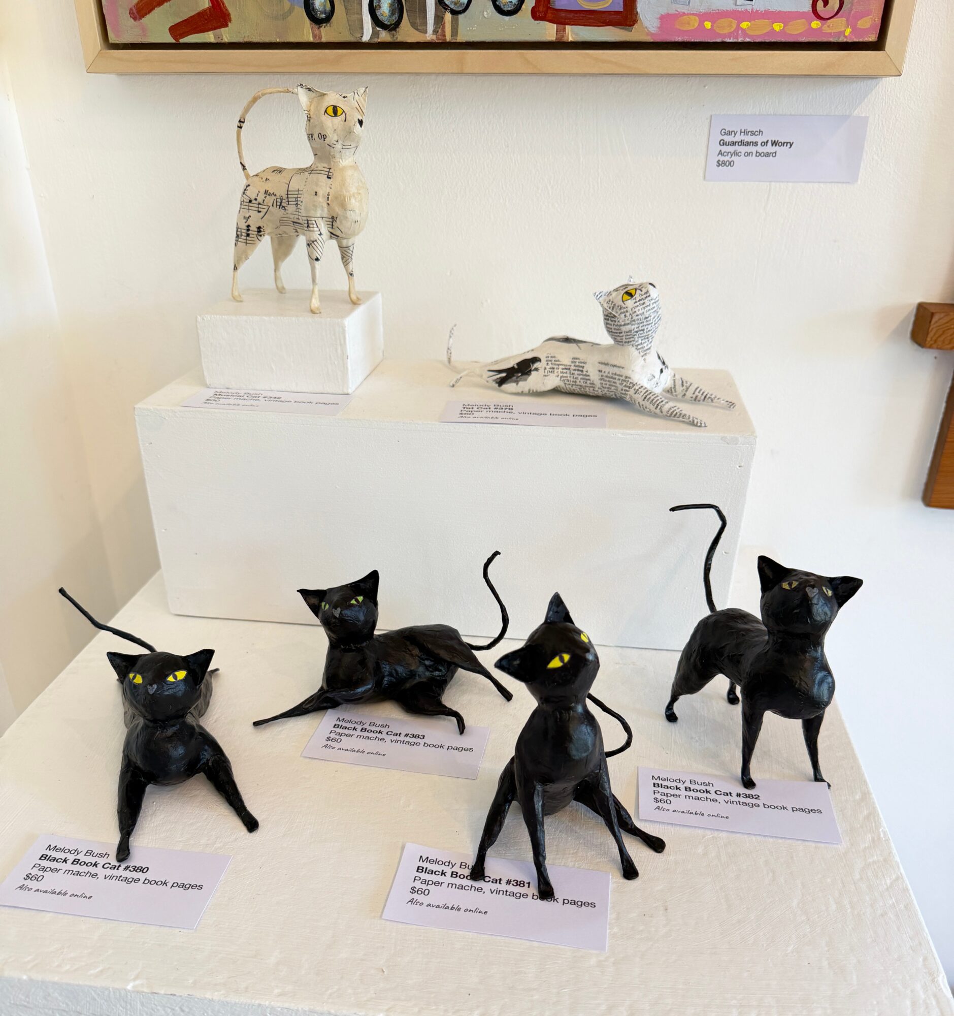 Five black cat sculptures with yellow eyes displayed on a table, with two animal figures on a white platform behind them.