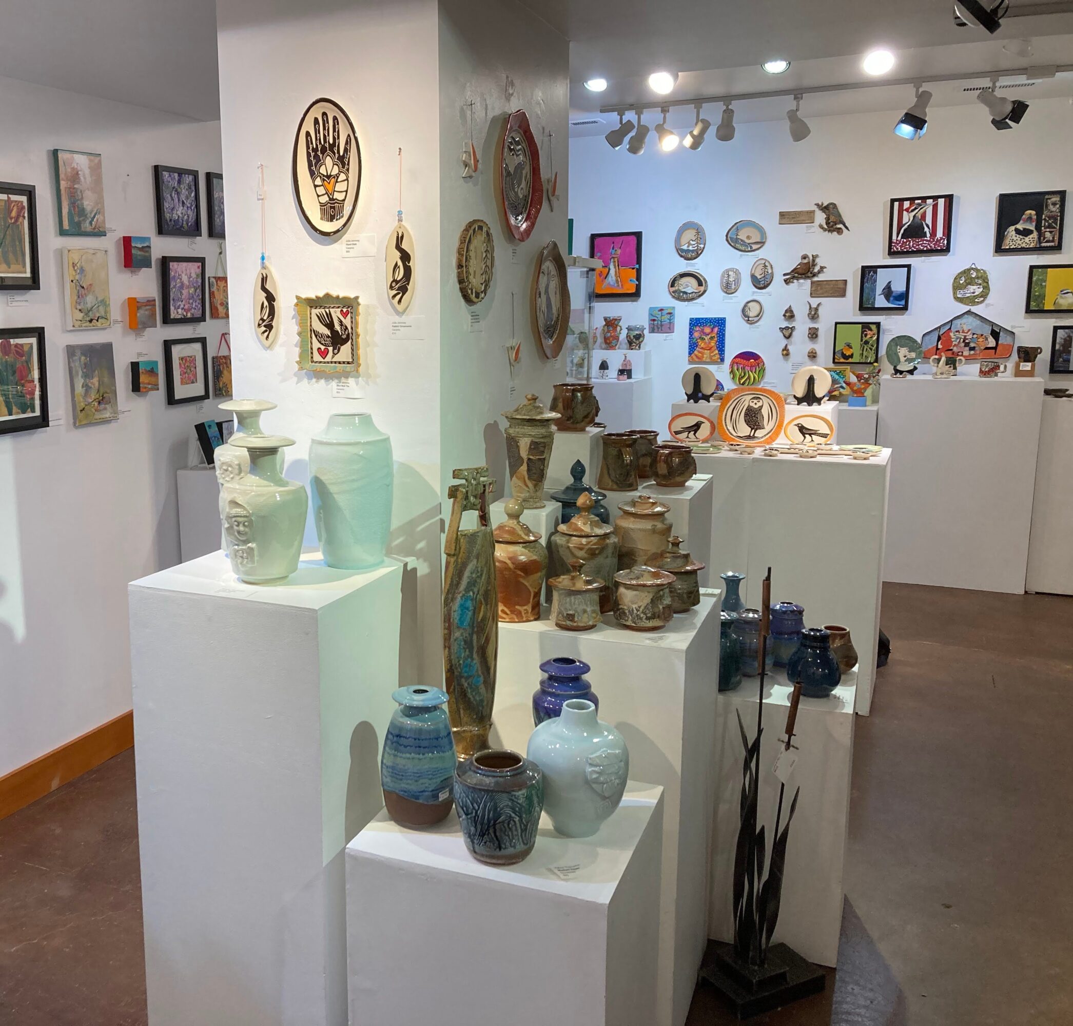 Art gallery with pottery and wall art displayed on white pedestals and walls, with paintings and decorative items in background.