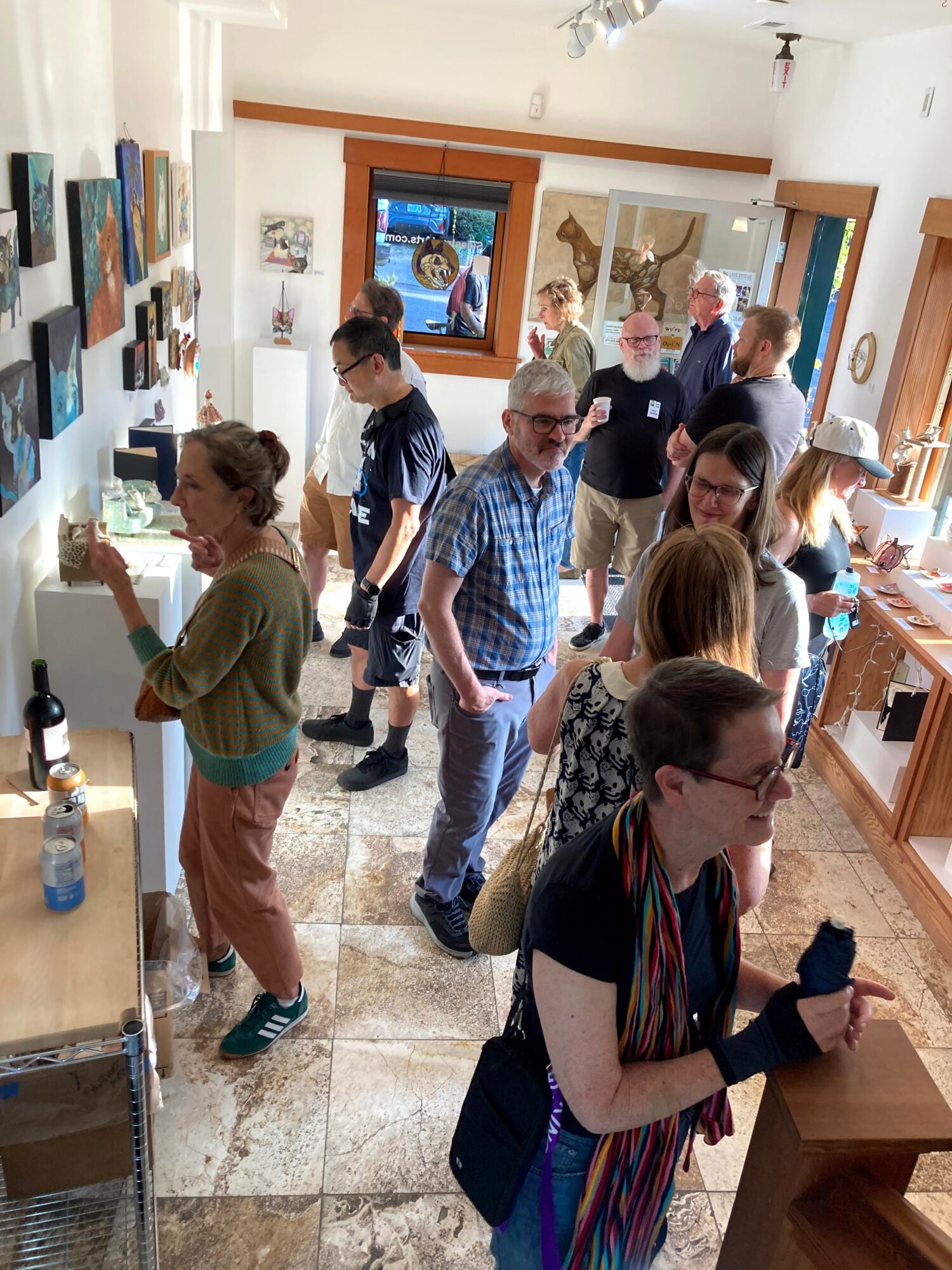 Group of people browsing art in a gallery, some looking at paintings on walls, others examining items on display, with sunlight coming through windows.