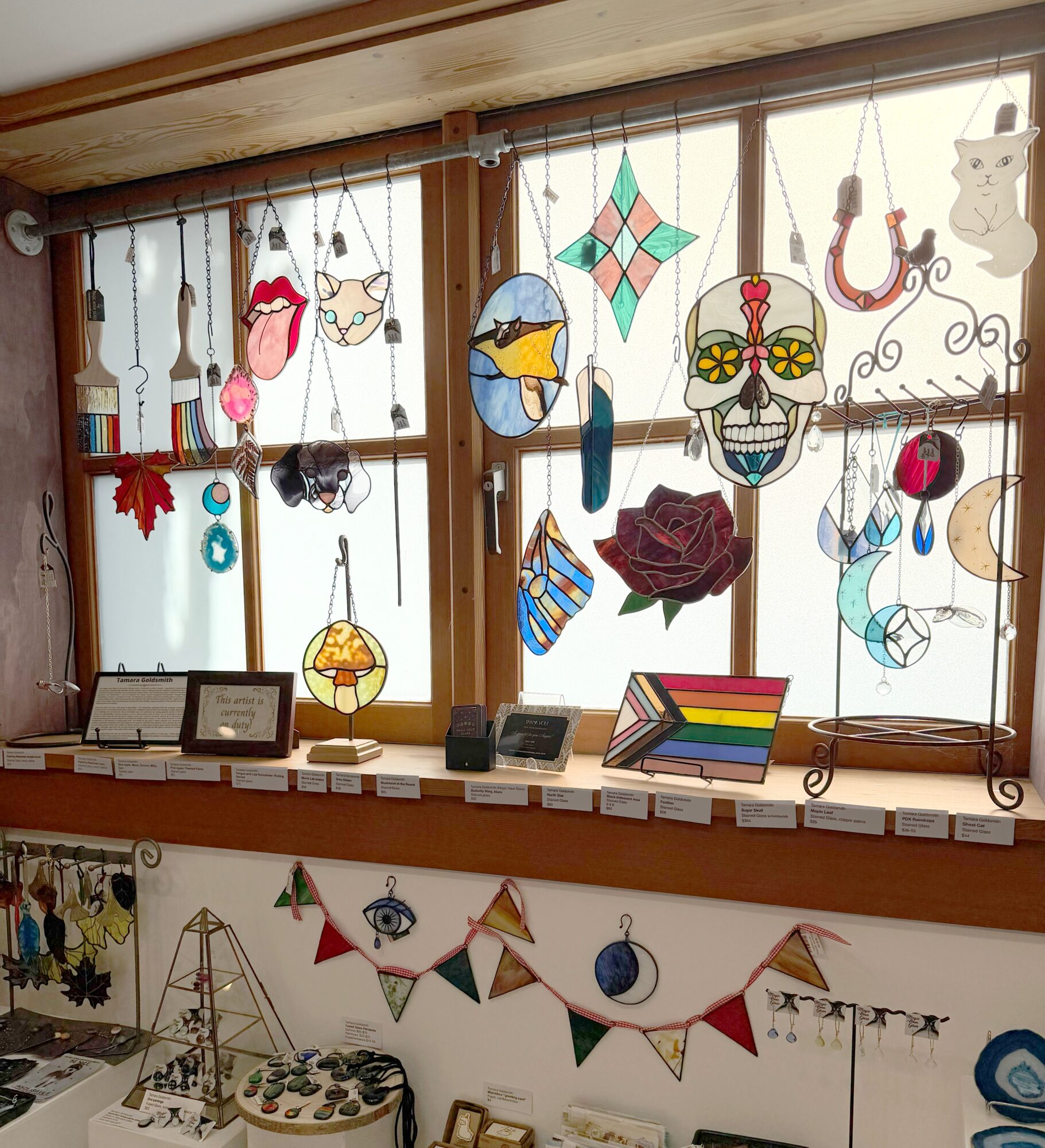 Decorative stained glass hanging ornaments on window, including a skull, cat, fish, and geometric shapes, with a display of jewelry below.