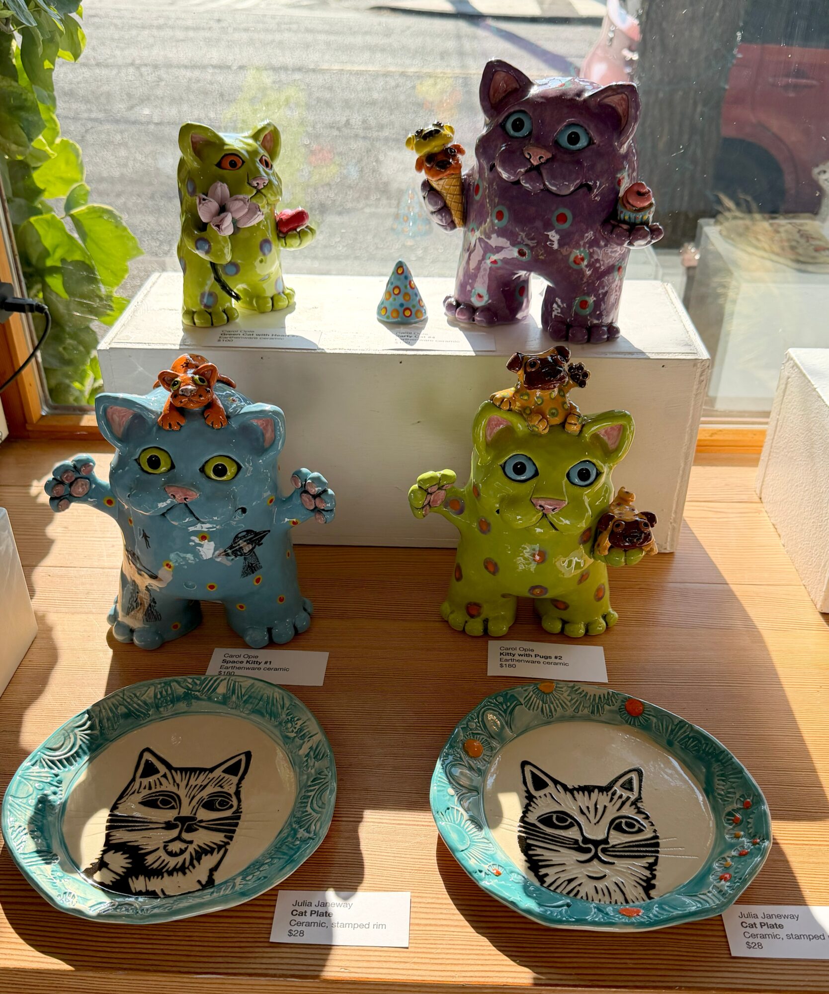 Six colorful cat figurines on a windowsill and table, with two decorative plates featuring cat illustrations, in a bright room.