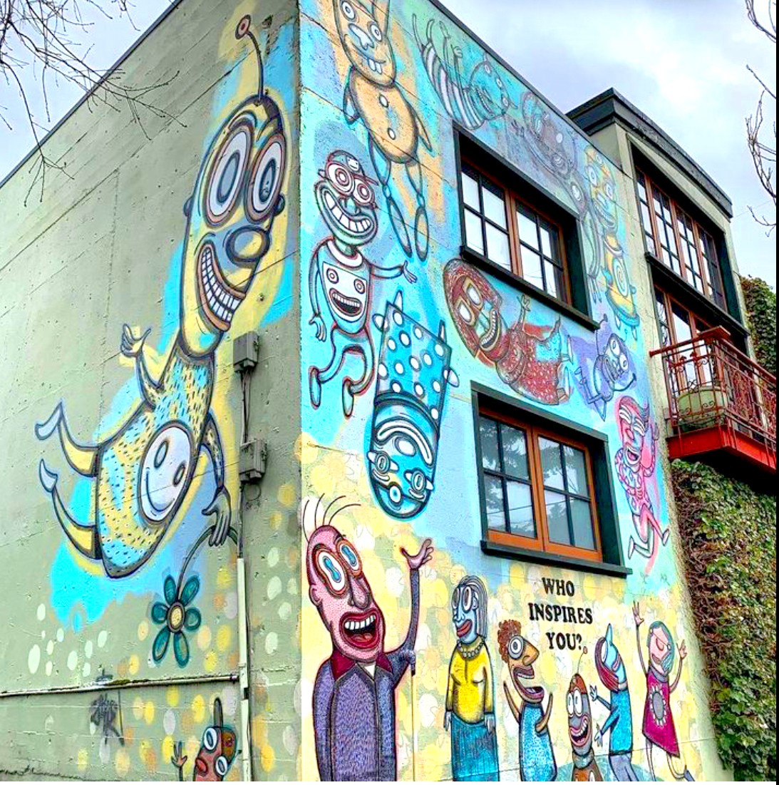 Colorful graffiti of cartoon characters on building corner, with a sign reading 'Who inspires you?' and windows above.