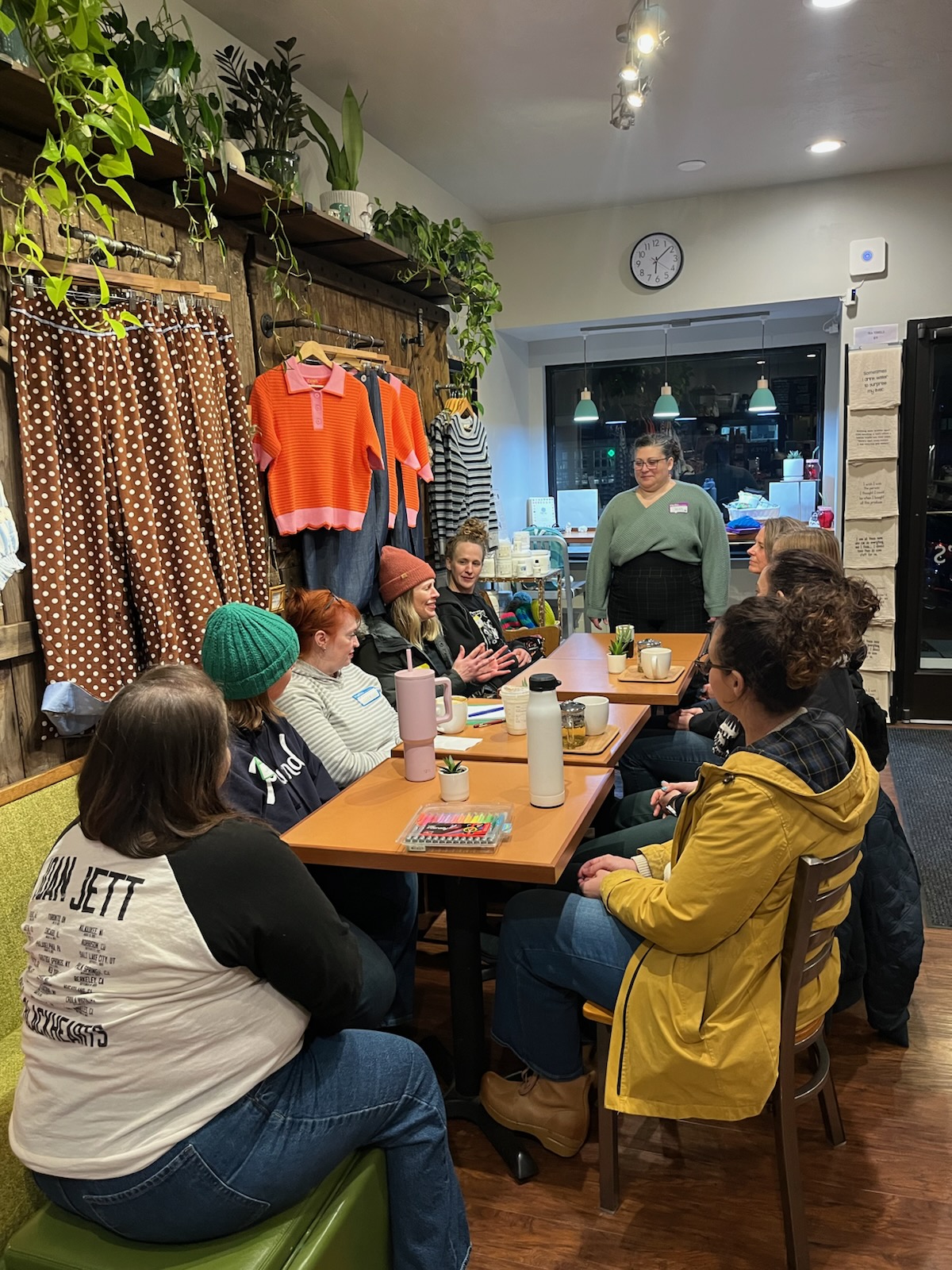 Group of people sitting around a table in a cozy room, with a person standing and speaking, plants and curtains in the background.