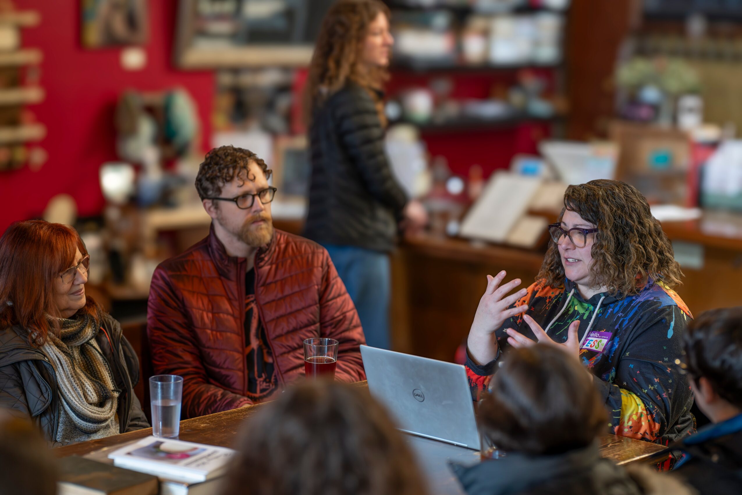 Group of people sitting at a table in a cozy cafe, engaging in conversation with a woman gesturing with her hands.