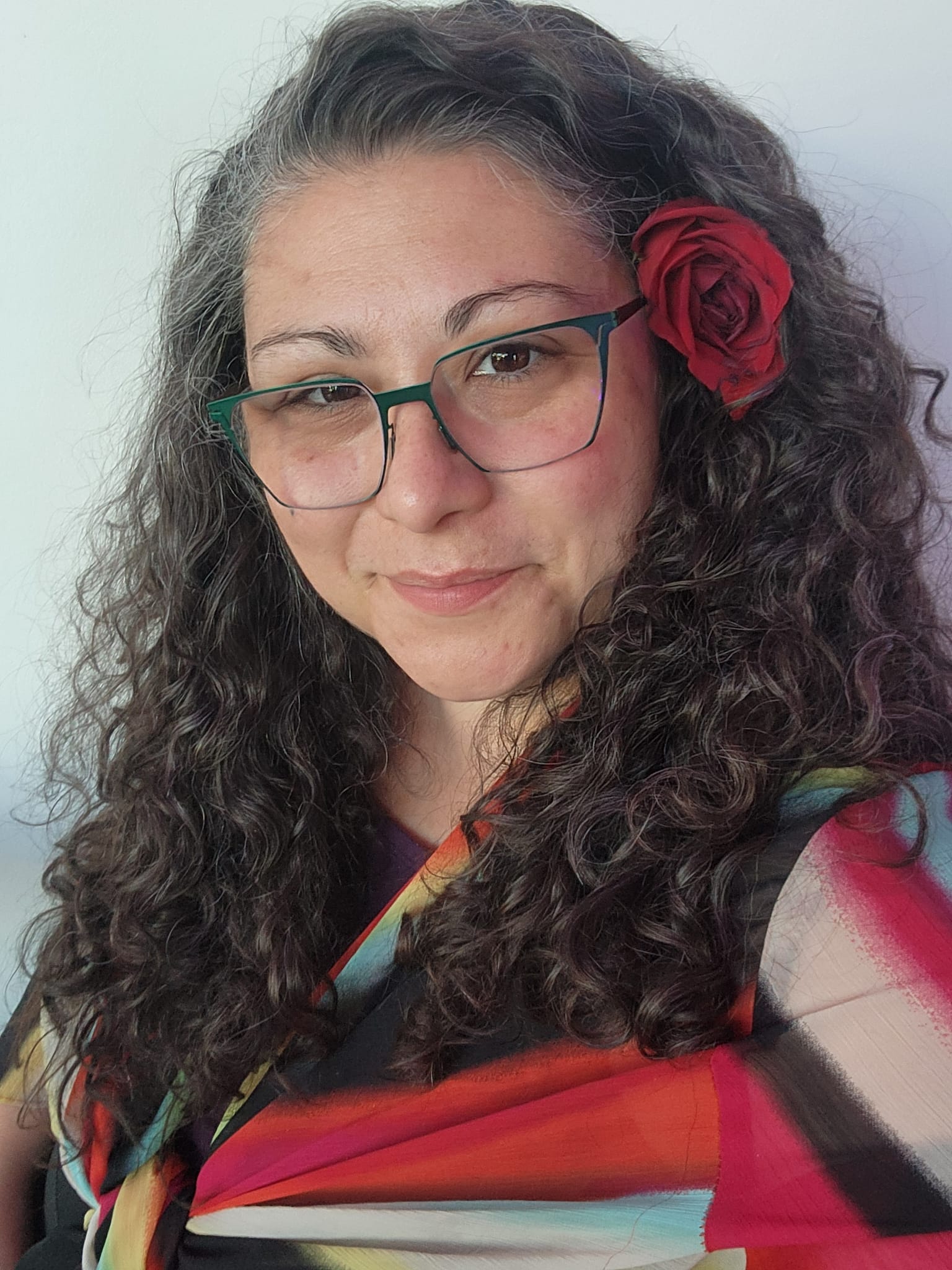 Woman with curly hair, glasses, and a red flower in her hair, smiling slightly, wearing a colorful top.