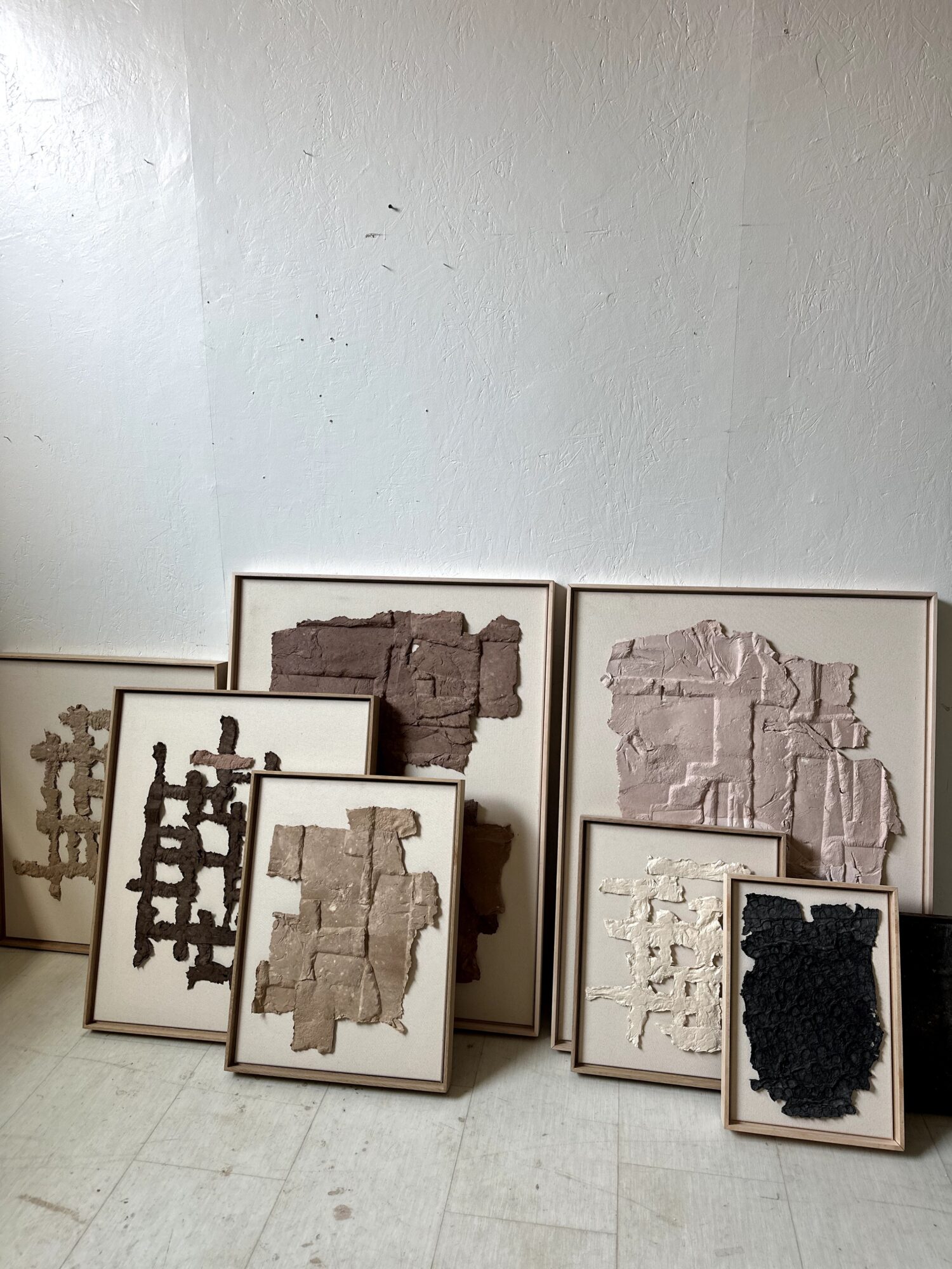 Collection of framed artwork with textured, abstract designs in various shades, leaning against a plain wall.