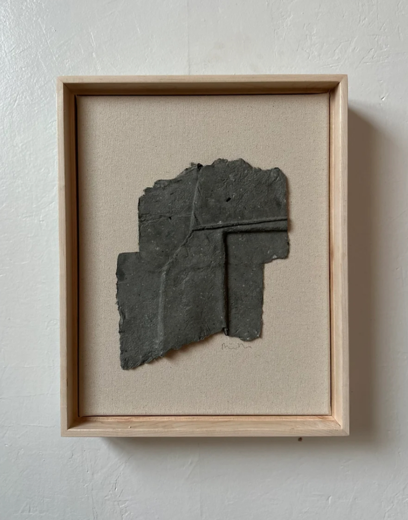 Fragment of dark gray stone with carved lines, mounted in a wooden frame with beige background.