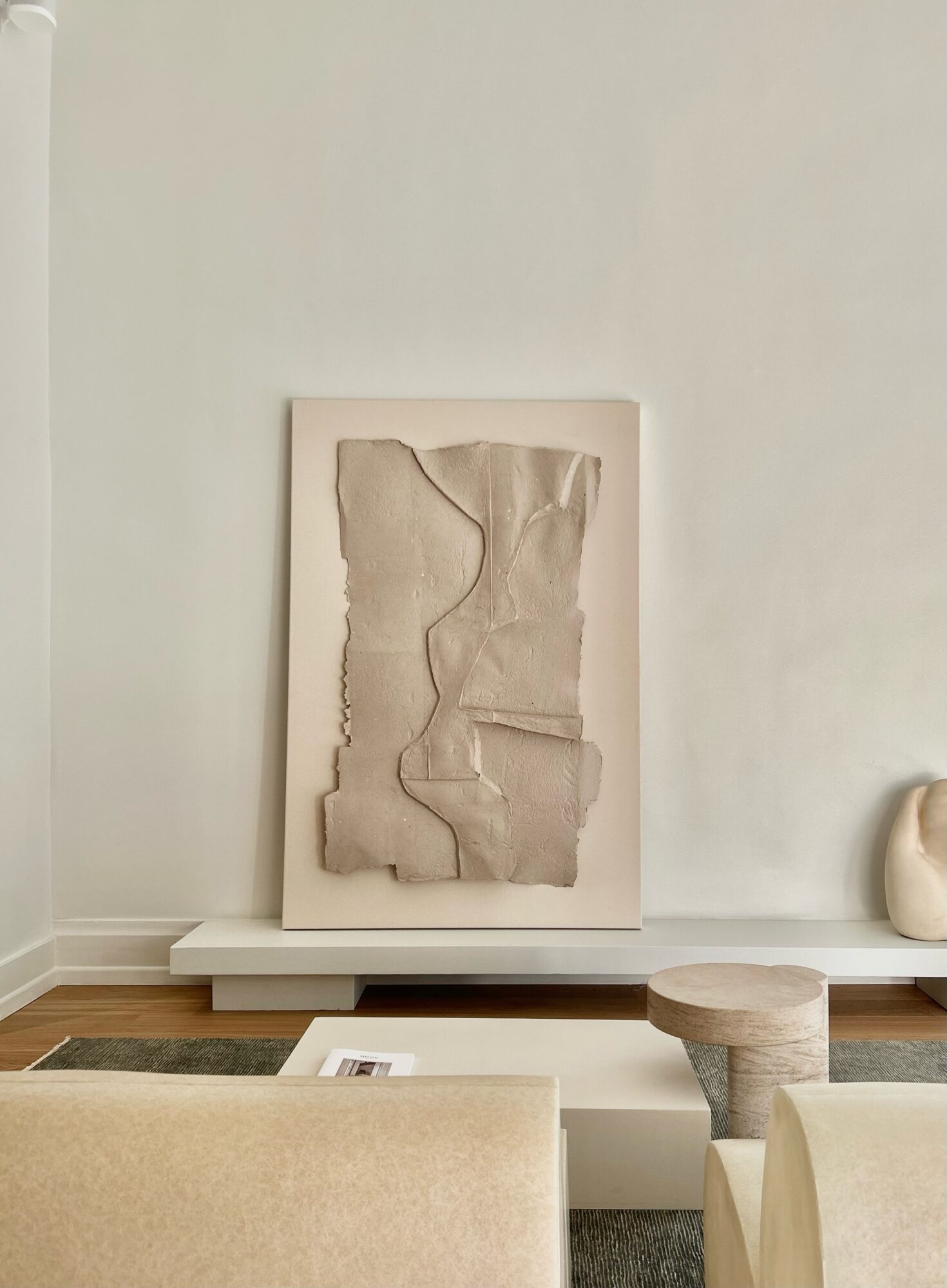 Abstract artwork with textured beige material on a white wall, with a small round stool and a beige chair in a modern room.