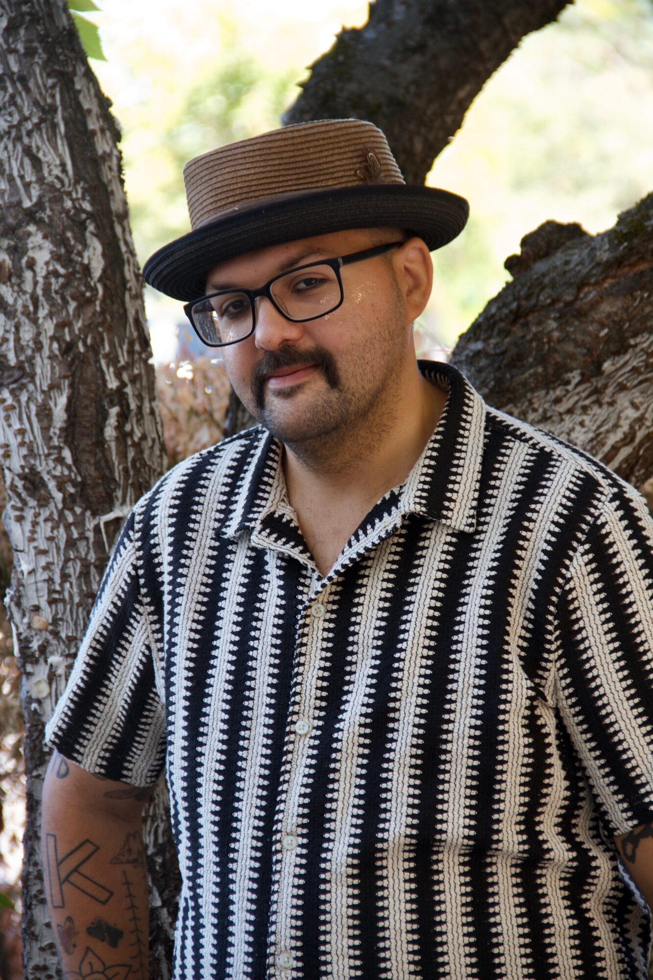 Man with glasses and a hat, wearing a patterned shirt, standing outdoors near tree trunks.