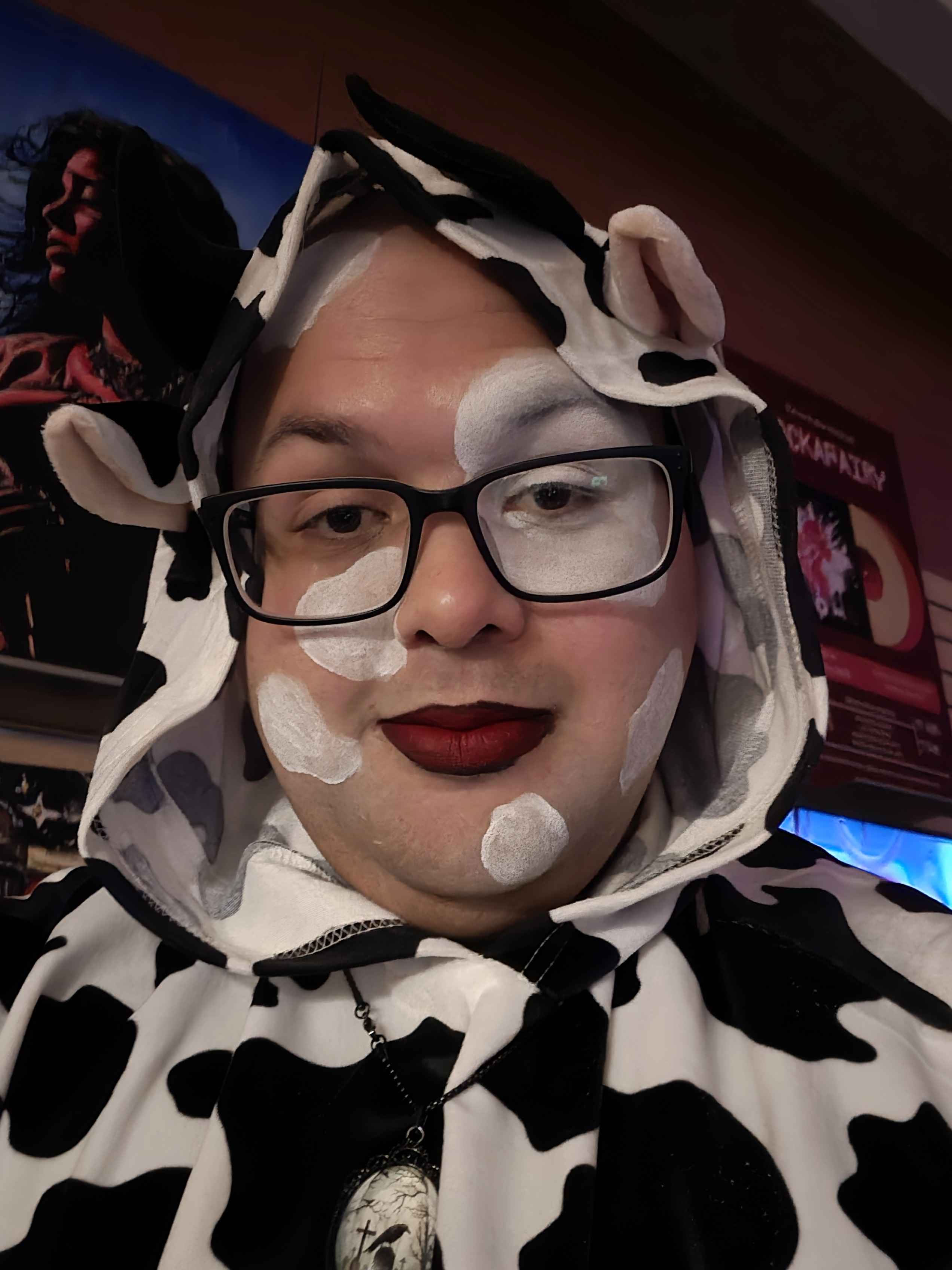 Person with makeup and glasses wearing a cow-patterned hoodie, in a dimly lit indoor setting.