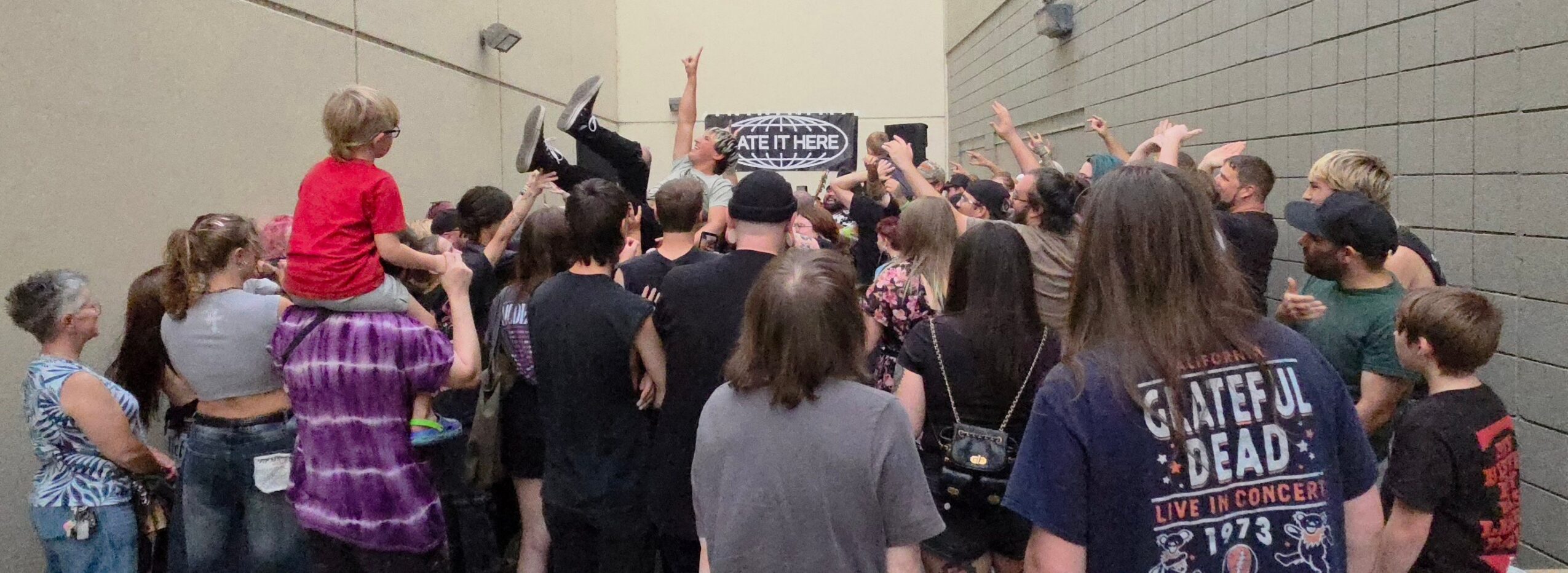 Group of people gathered outdoors, some raising hands, with a sign in the background, in a casual setting.
