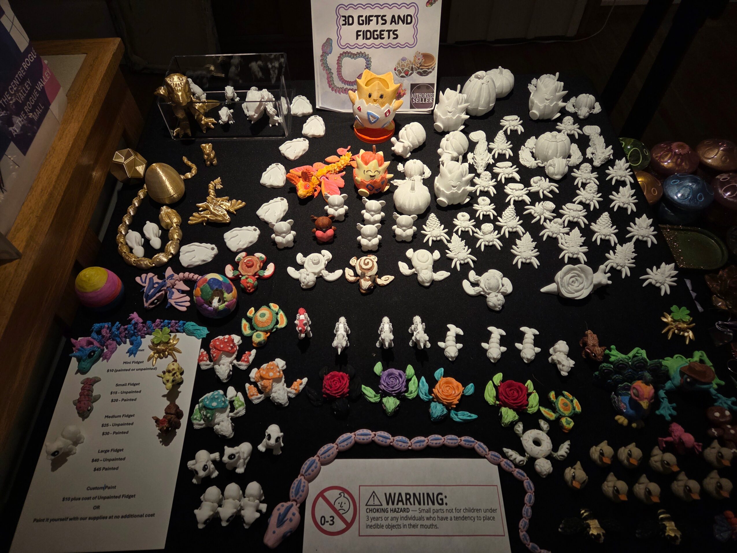 Display of small toys, figurines, and jewelry on a black table, with a sign and a paper at the bottom left.
